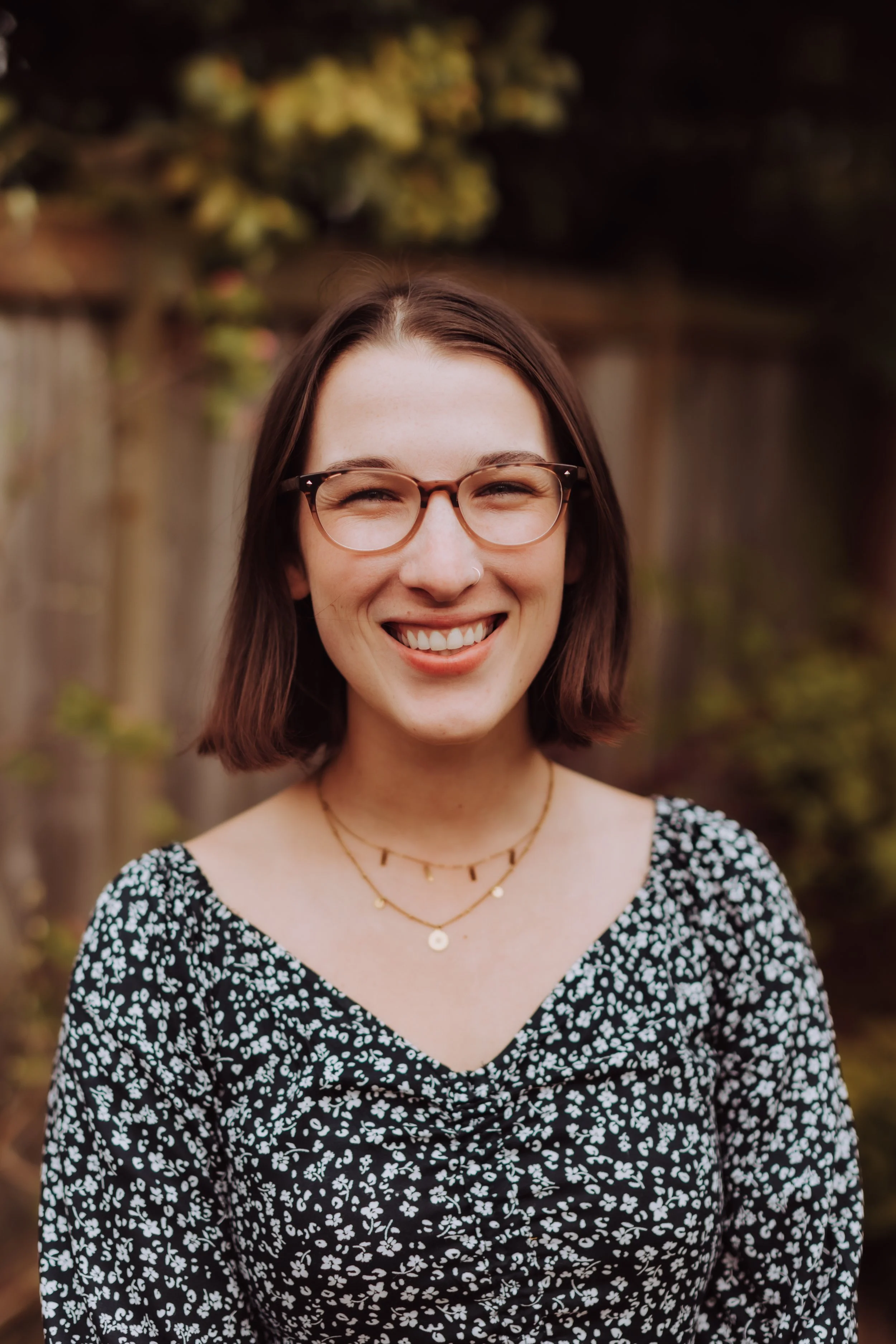 A young woman with brown hair, wearing glasses, smiling outdoors. She has a septum piercing and is wearing layered gold necklaces and a black dress with a white floral pattern. The background contains a wooden fence and greenery.