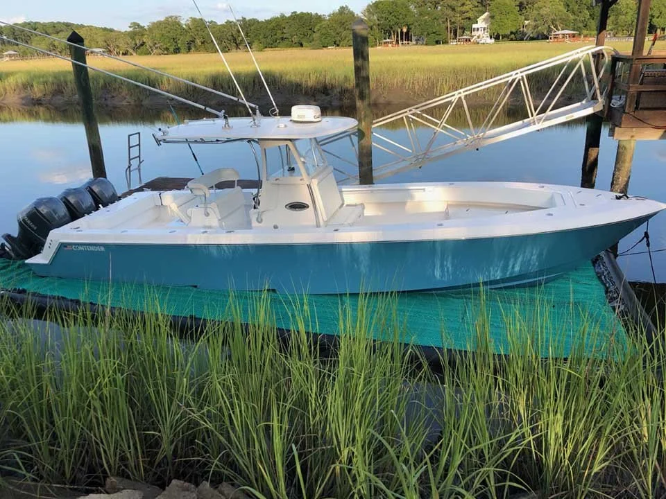 White and teal boat docked on a green mat next to tall grass and calm water with a small wooden dock and pole support in the background.