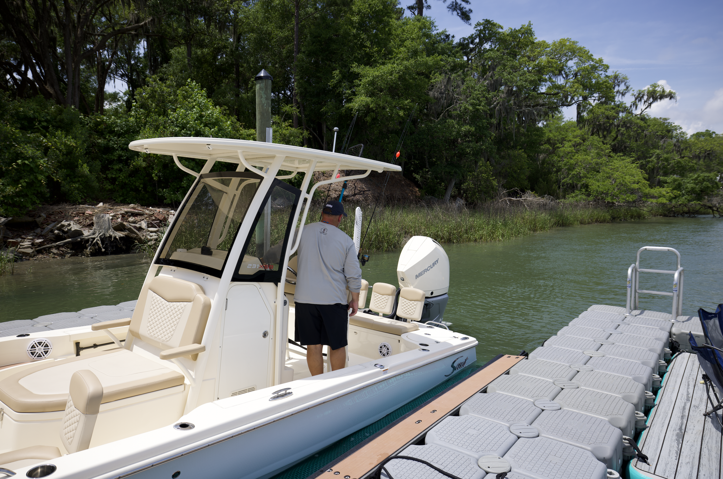 A man standing on a white boat docked in water near a wooded shoreline, with an attached floating dock and multiple gray storage containers.