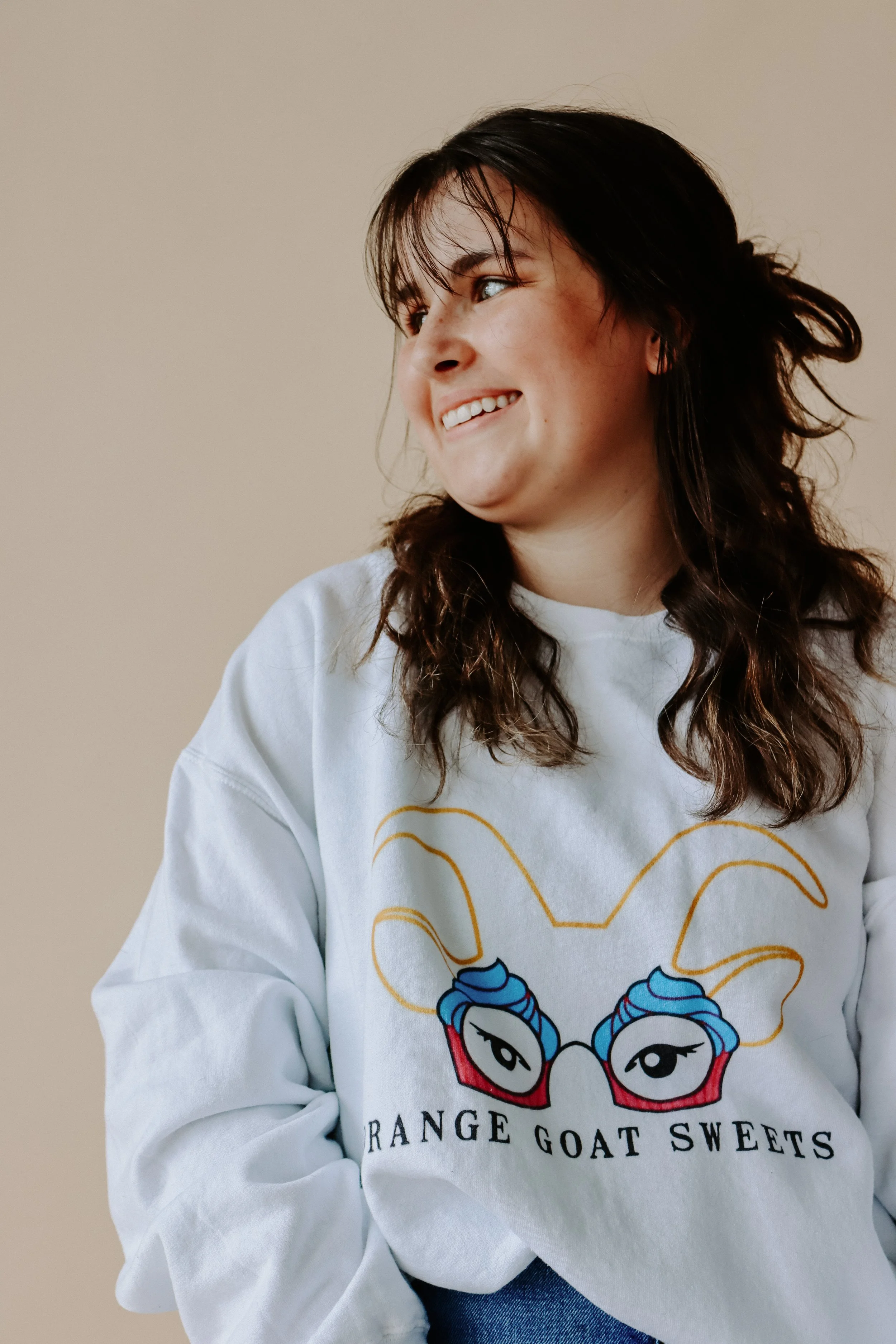 A woman with dark, wavy hair smiling and looking to the left, wearing a white sweatshirt with an illustrated goat face with blue hair and red glasses, and the words "Range Goat Sweets" on it.