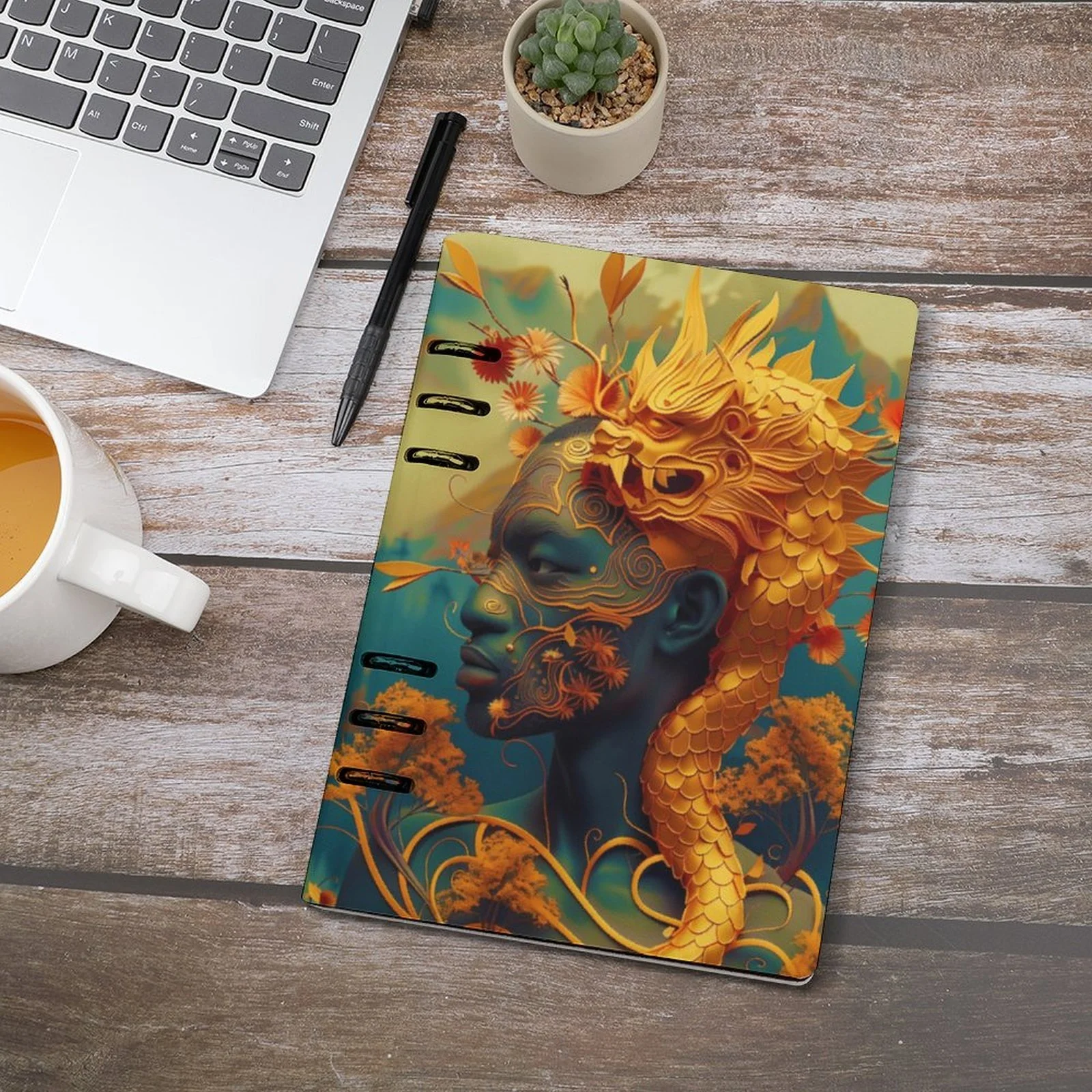 A tabletop with a laptop, a cup of coffee, a pen, a small potted succulent, and a journal featuring a vibrant cover design of a fantastical figure with ornate patterns and a dragon motif.