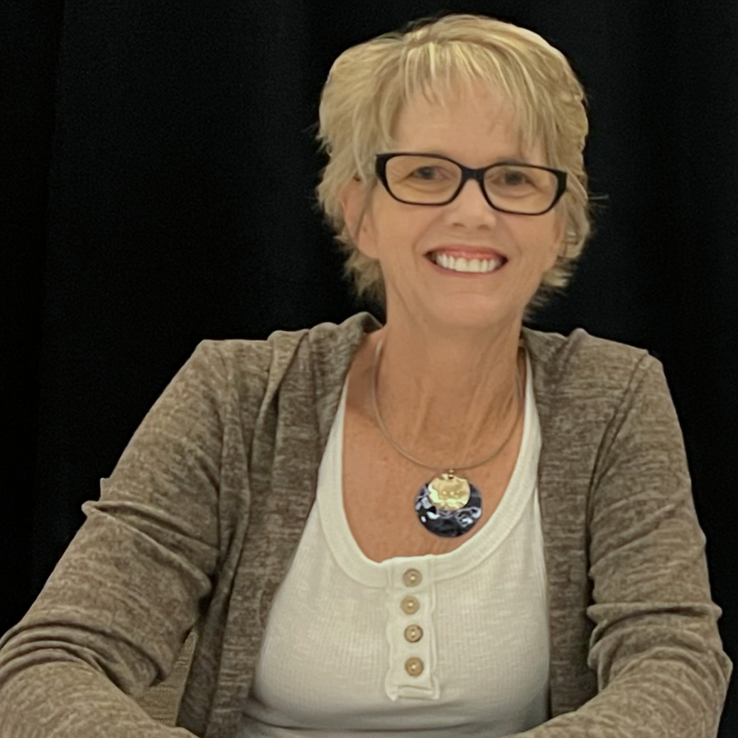 A smiling woman with short blonde hair, glasses, and a necklace, wearing a beige cardigan over a white top, sitting against a black background.