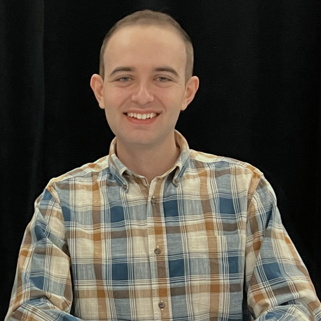 A young man with short hair, wearing a plaid button-up shirt, smiling against a black background.