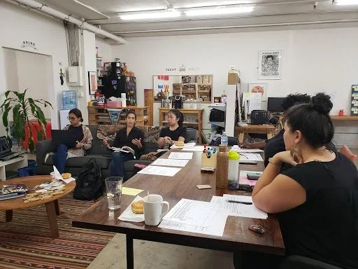 Five women in an office, sitting at desks or on a couch working on laptops