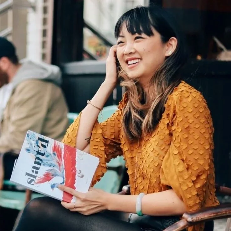 Krystie Yen, Slant'd Founder and Executive Director pictured smiling at a cafe with a Slant'd Magazine in hand.