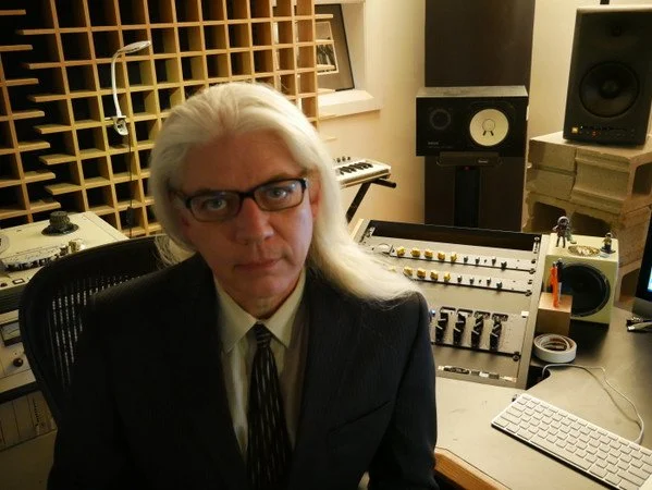 Man with long white hair and glasses in a recording studio, surrounded by audio equipment, mixers, and speakers.
