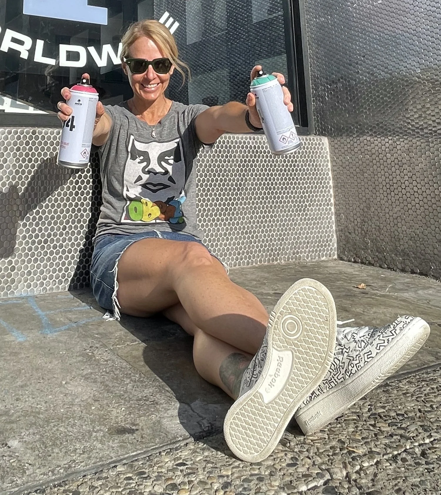 Smiling woman with sunglasses holding two spray paint cans, sitting on a tiled surface wearing a graphic t-shirt, denim shorts, and patterned sneakers.