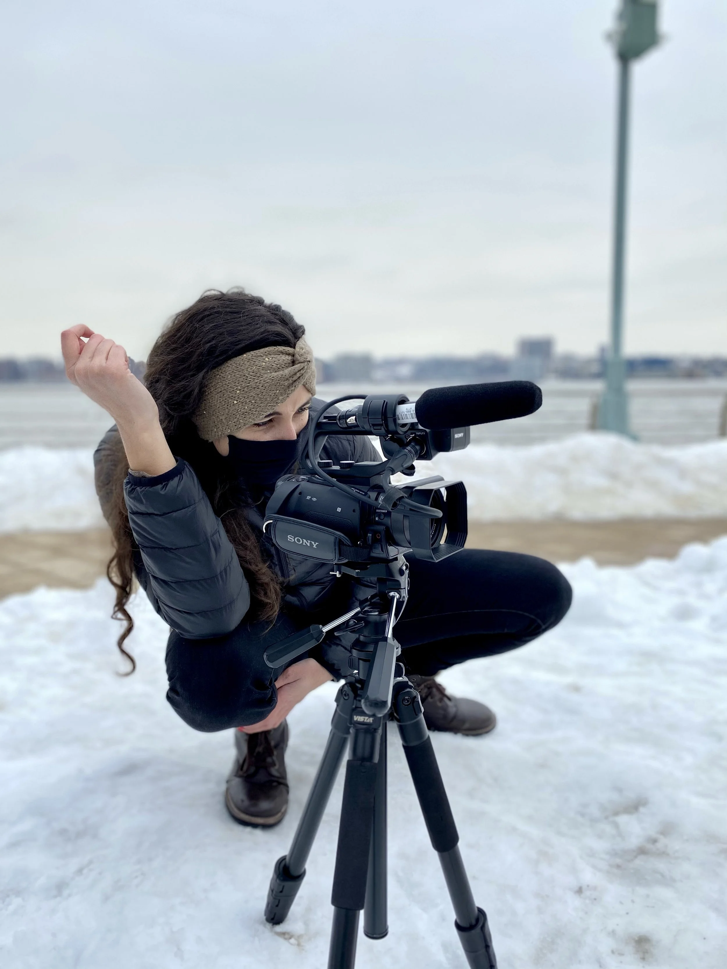 Person wearing a headband, mask, and winter jacket operating a camera on a tripod in a snowy outdoor setting.