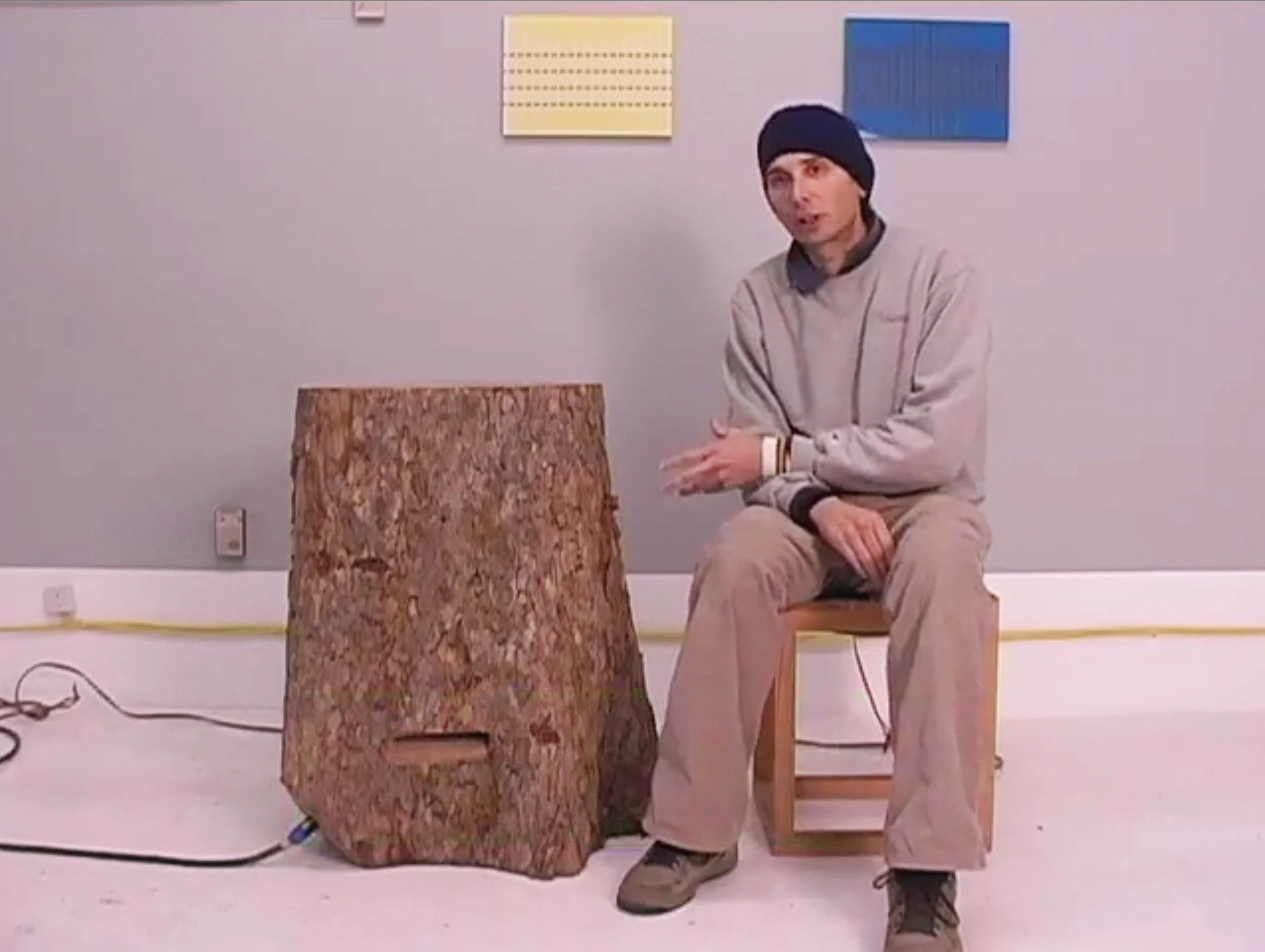 Person sitting on a stool next to a large tree stump indoors, with two small abstract art pieces on a gray wall behind.