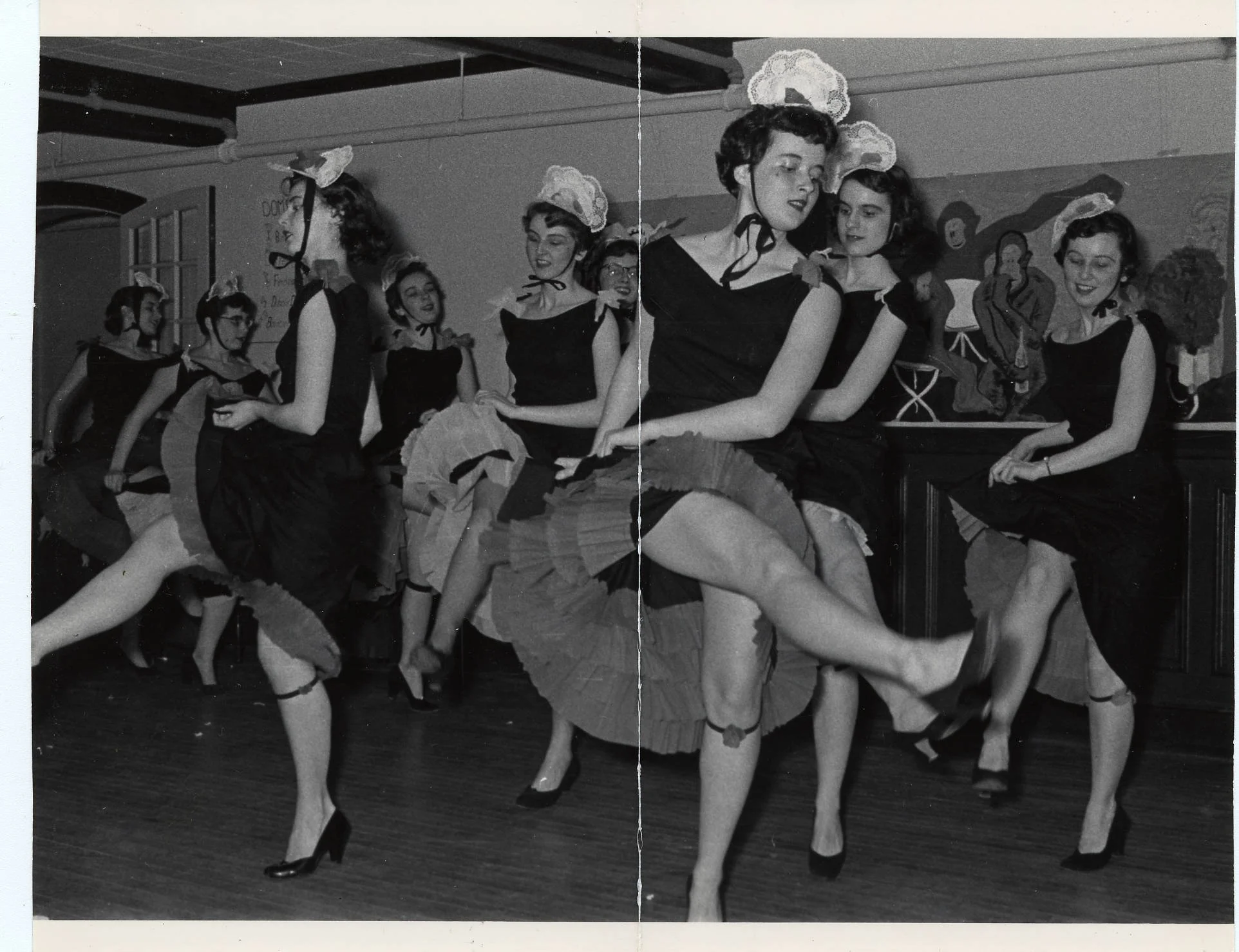 Group of women performing a can-can dance in a vintage setting, wearing black dresses, stockings, and headbands with lace details.