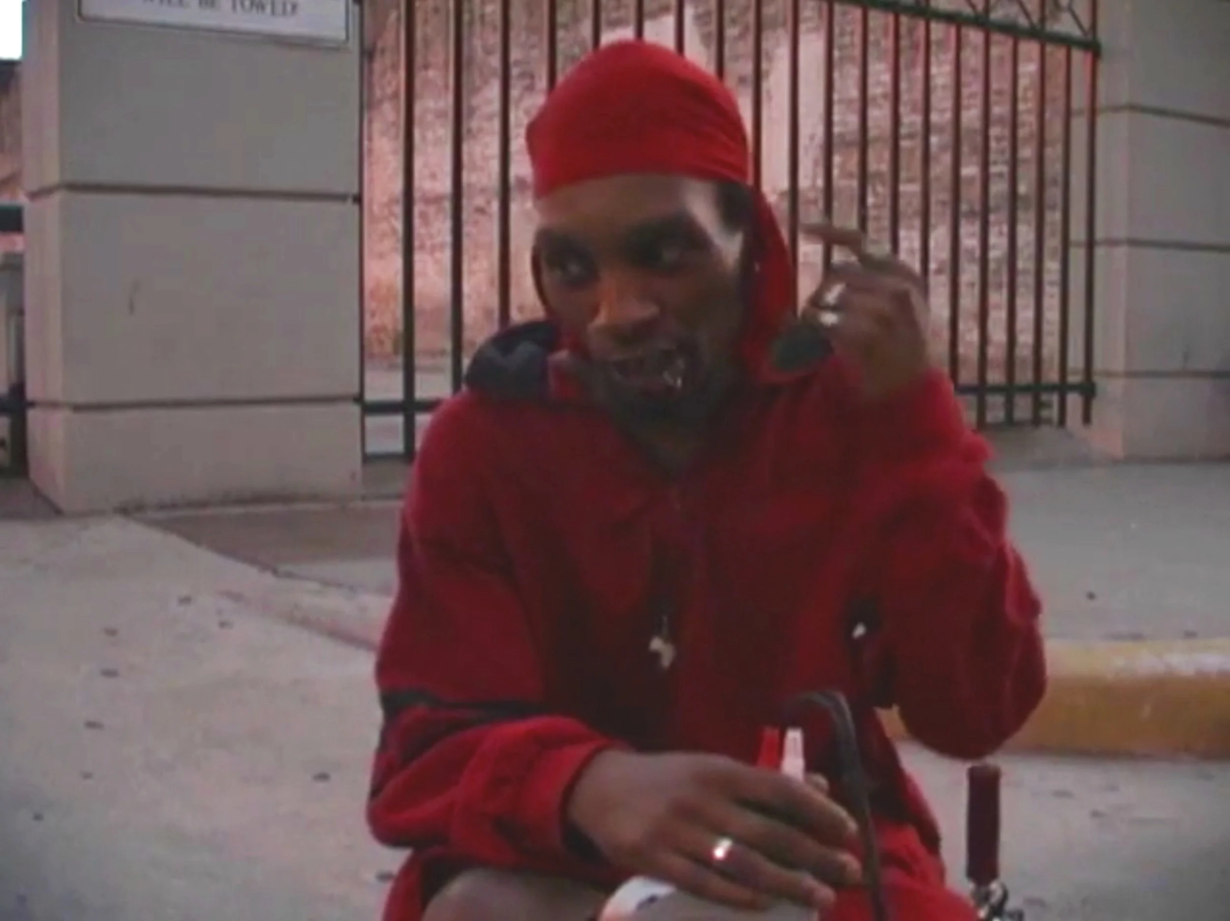 Person sitting outdoors wearing a red outfit and head covering, holding a small object in their hand, near a gated area.