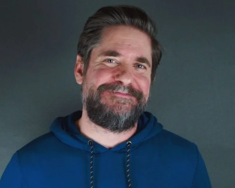 Man with a beard wearing a blue hoodie smiling against a gray background.