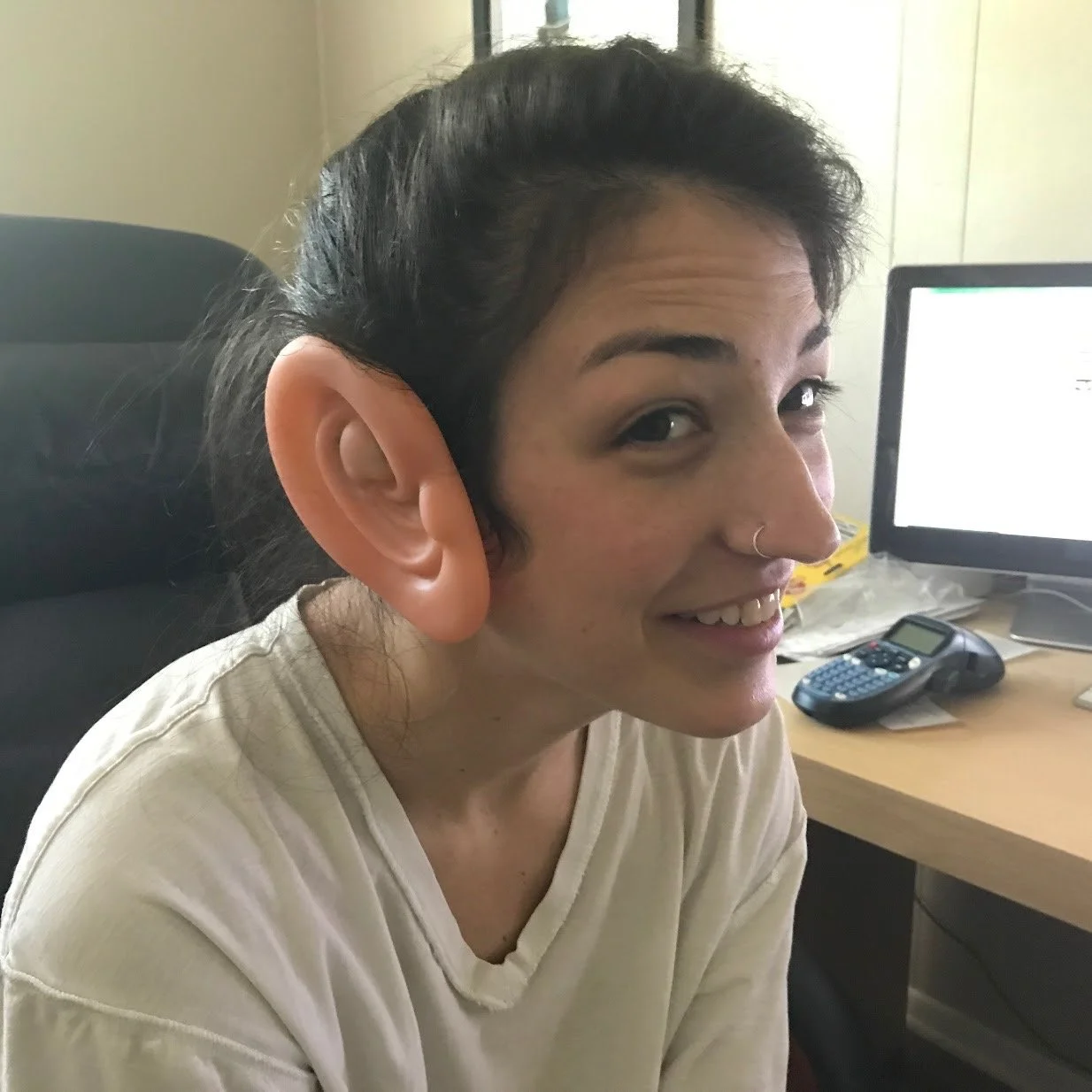 Person wearing oversized novelty ears, sitting at a desk with a computer in the background.
