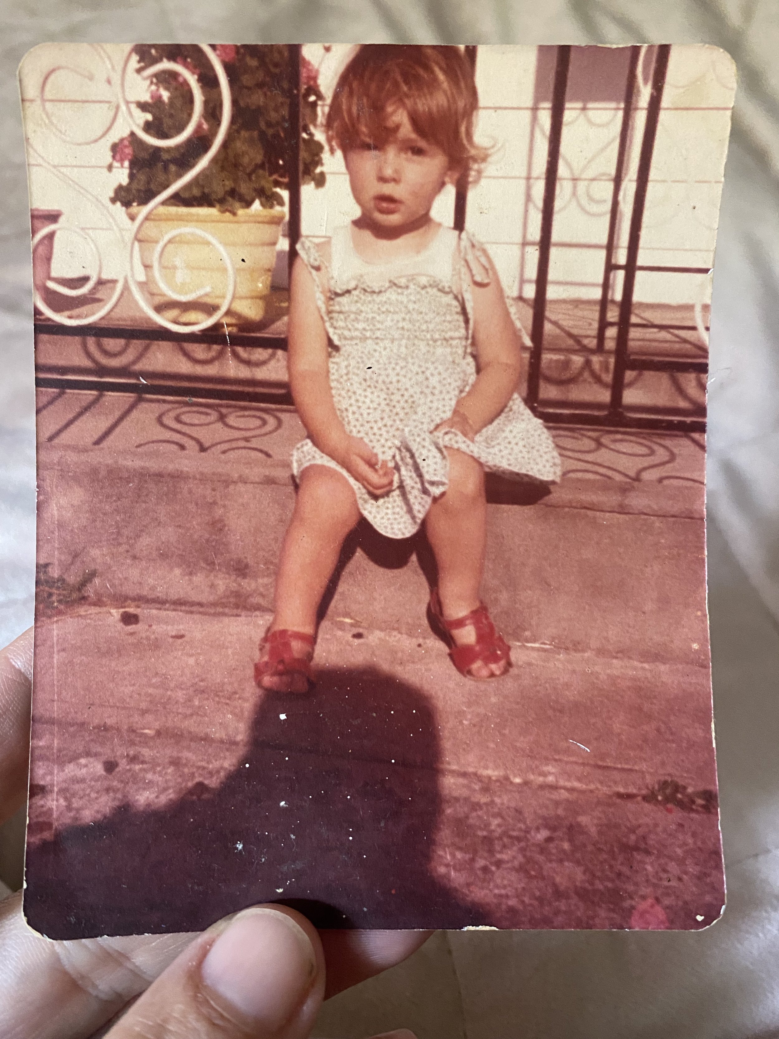 Nicole as a young child in a dress sitting on a concrete step in front of a metal railing with potted plants.