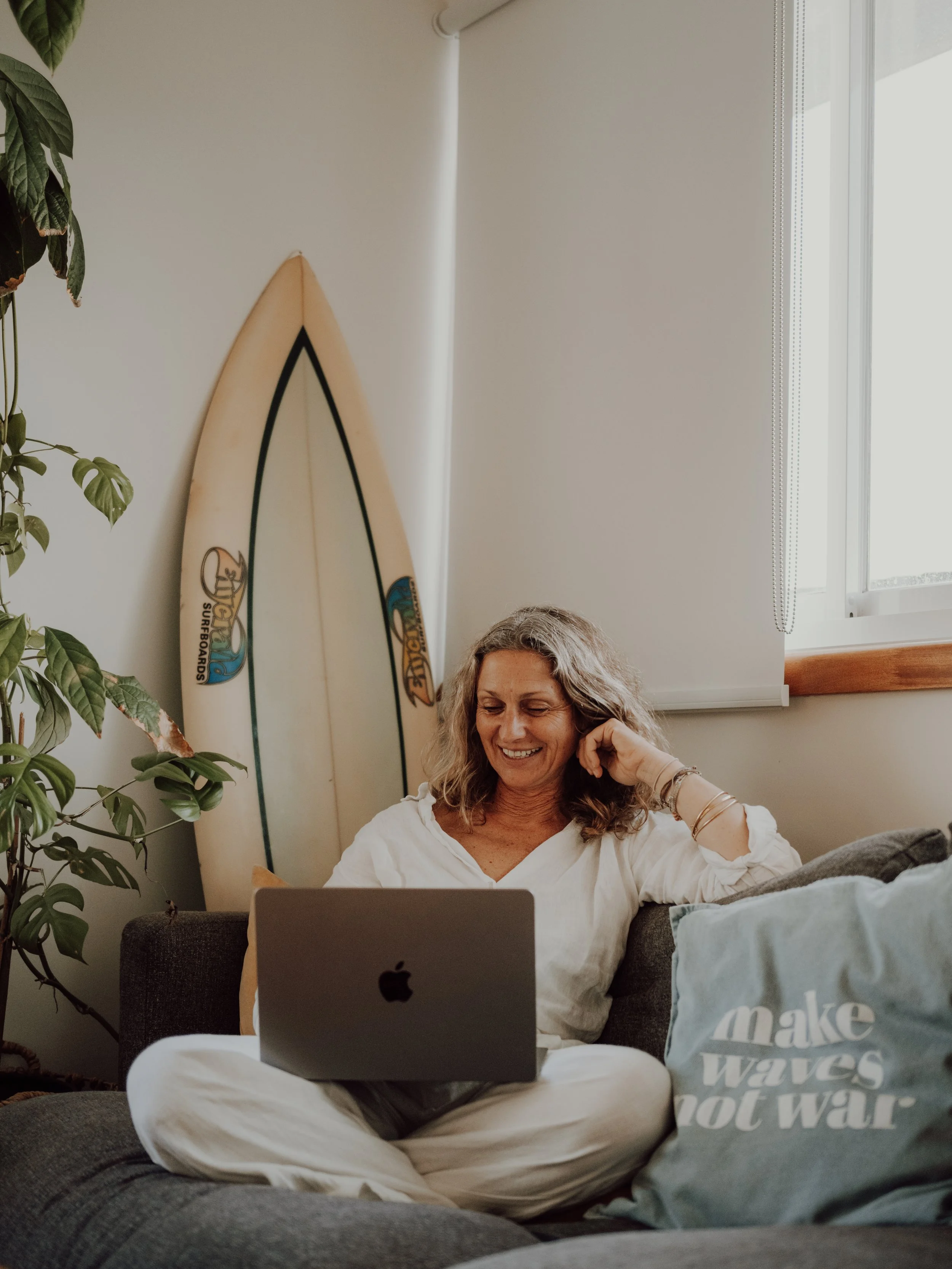 Nicole sitting on sofa with laptop, surfboard in background, pillow with 'make waves not war' text, houseplant nearby.