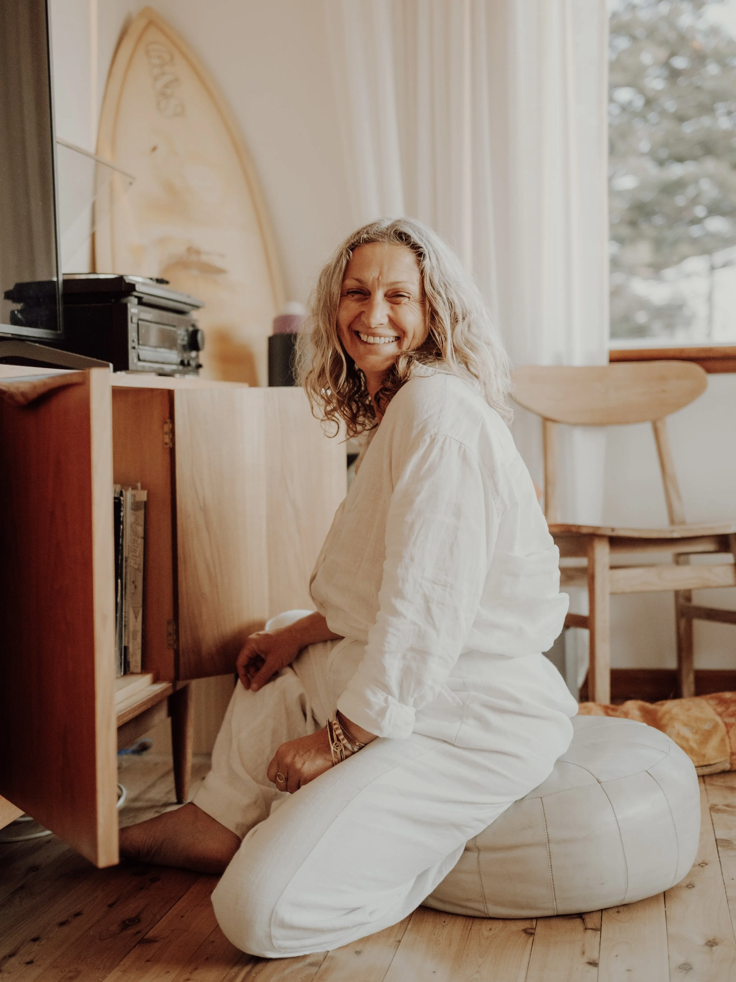 A smiling Nicole with her curly hair sitting on a cushion, wearing white clothing, in a cozy room with wooden furniture and a surfboard leaning against the wall.
