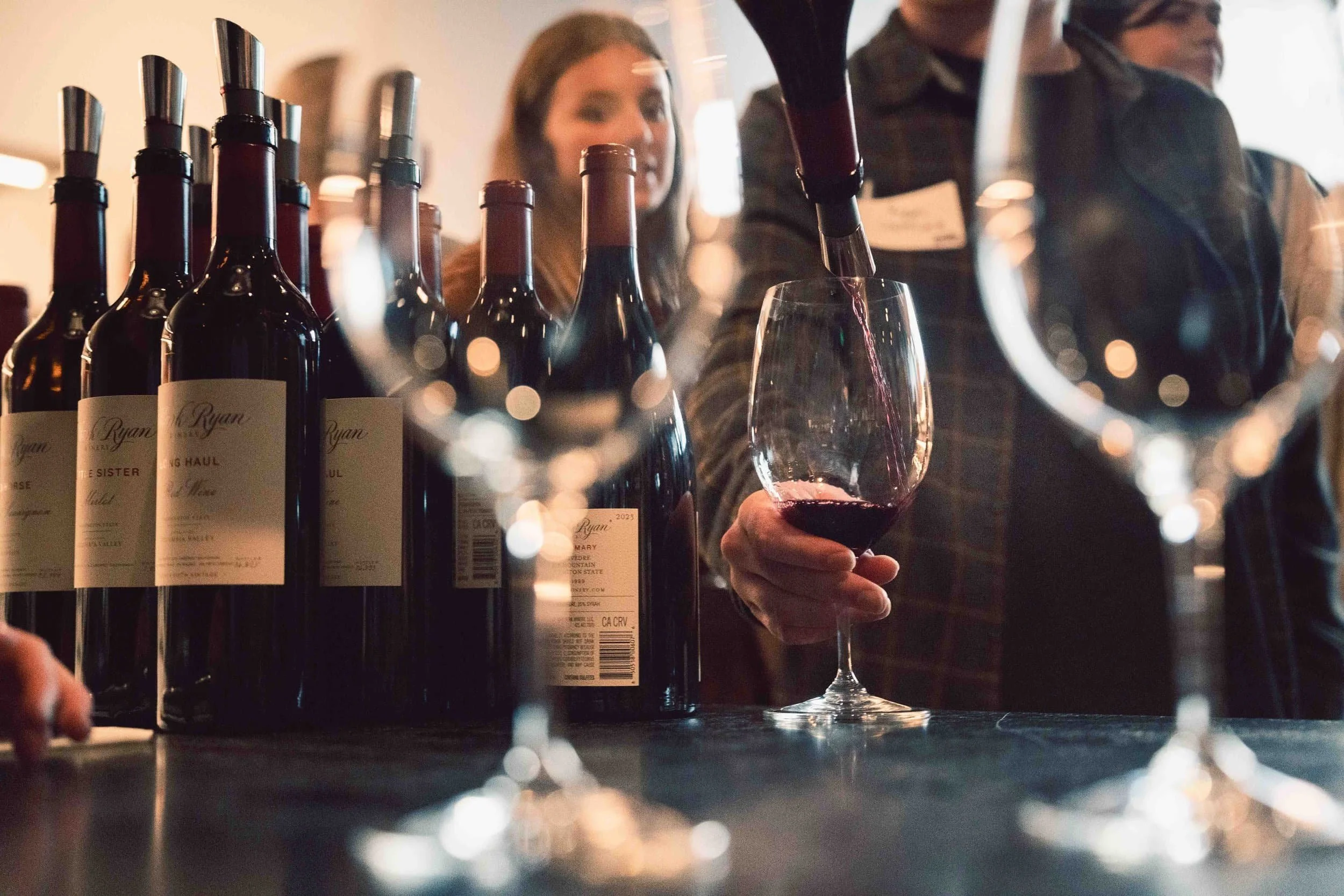 A glass of wine being poured on an metal counter, with a customer eagerly eyeing the glass.