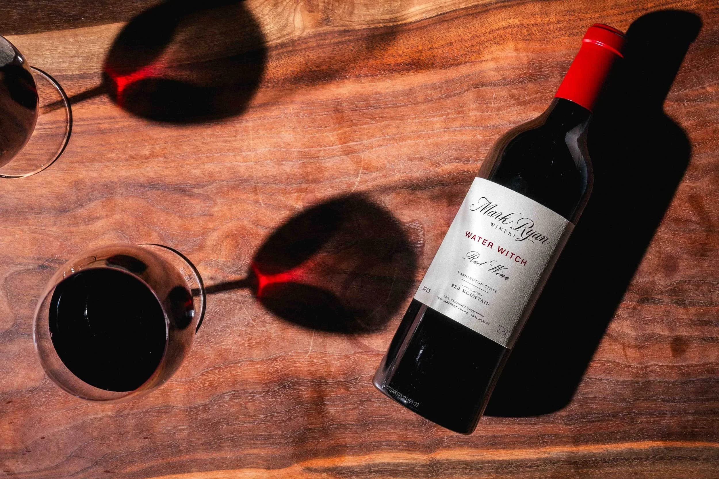 Bottle of Mark Ryan Water Witch Red Wine on a wooden table next to two wine glasses casting long shadows.