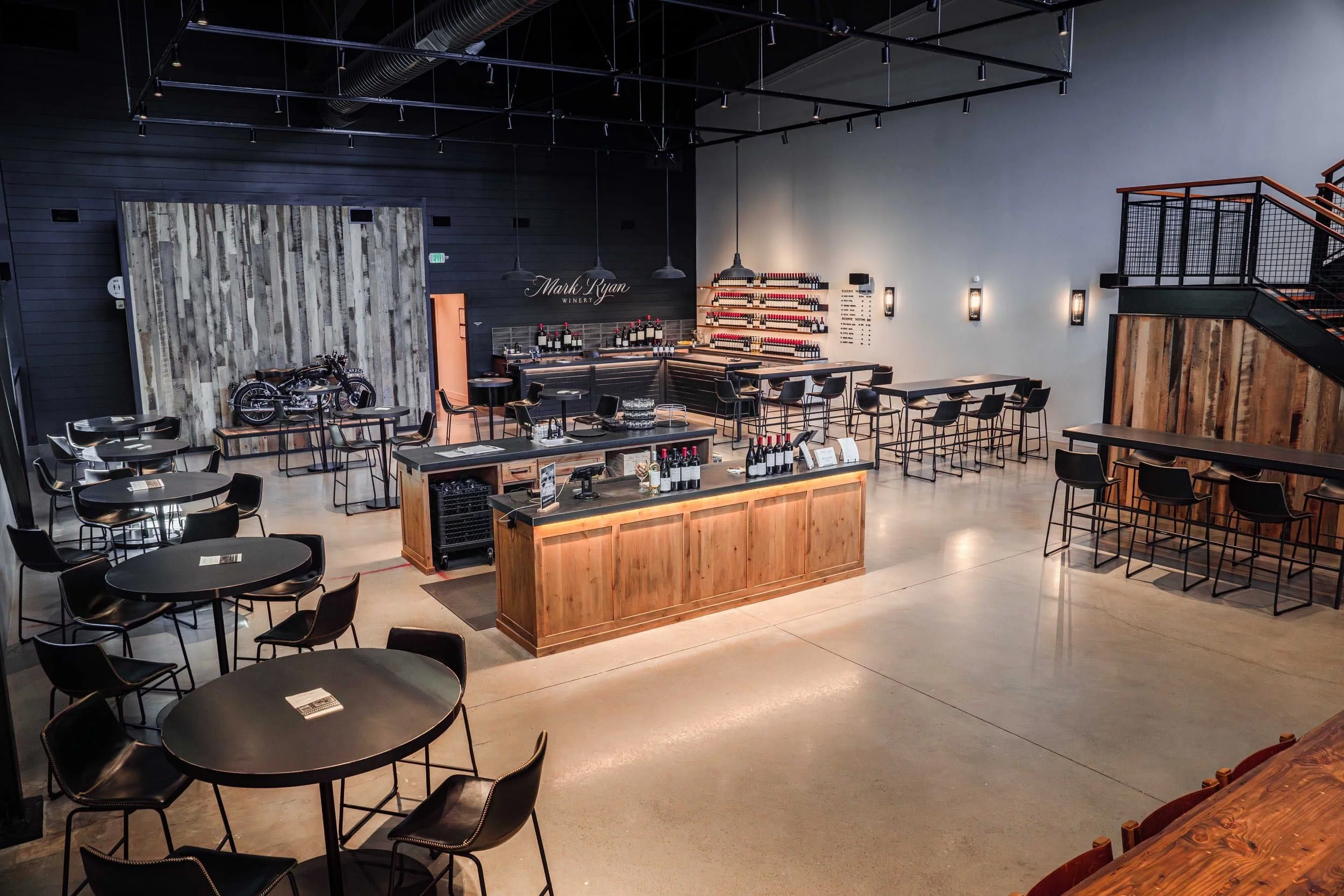Interior of the Mark Ryan Woodinville tasting room with modern black tables and chairs, a wooden bar with wine bottles, a vintage motorcycle display, and a staircase with wood and metal railing.