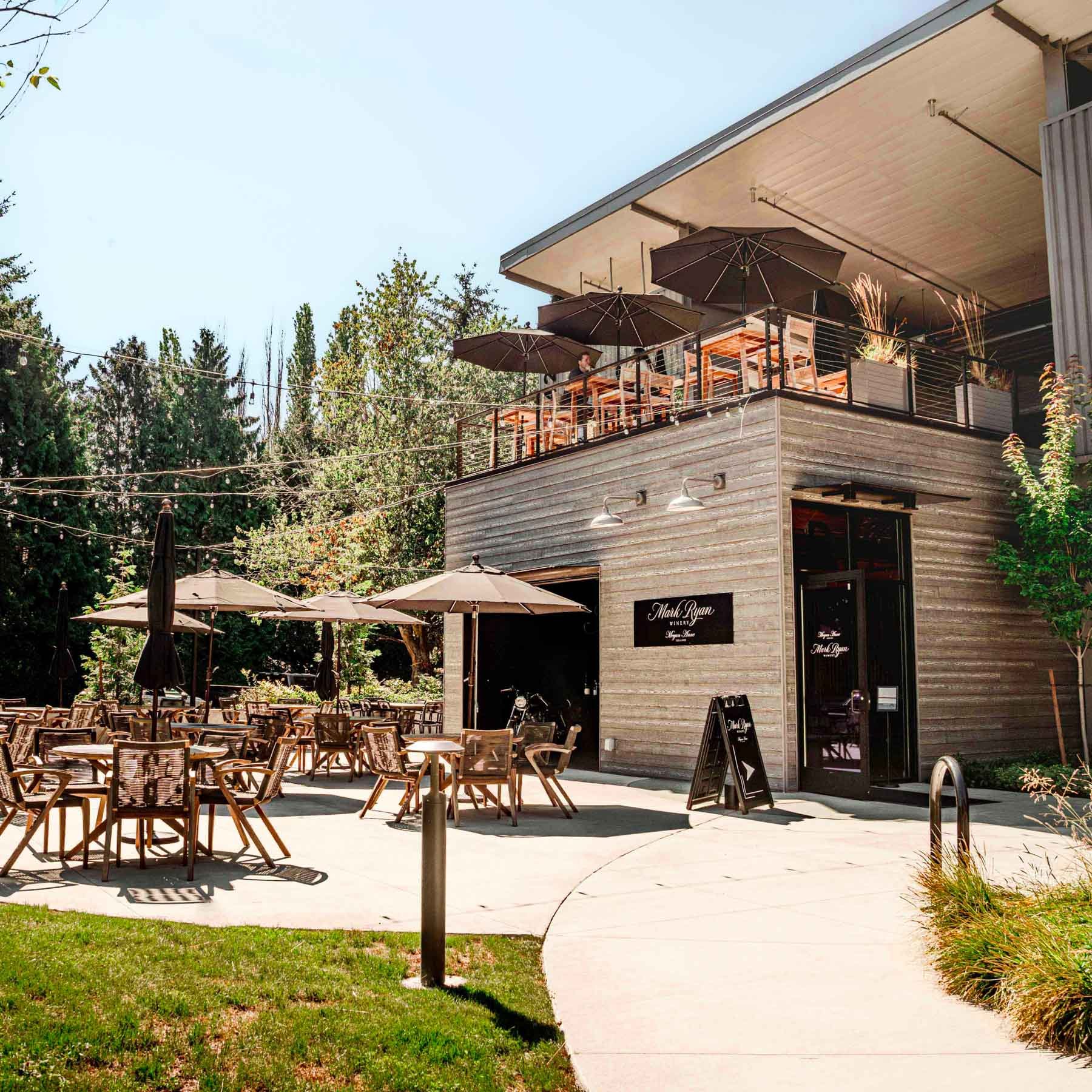 Street view of the Mark Ryan Winery Woodinville Tasting Room featuring a modern building with comfortable patio furniture and a deck lined with umbrellas.