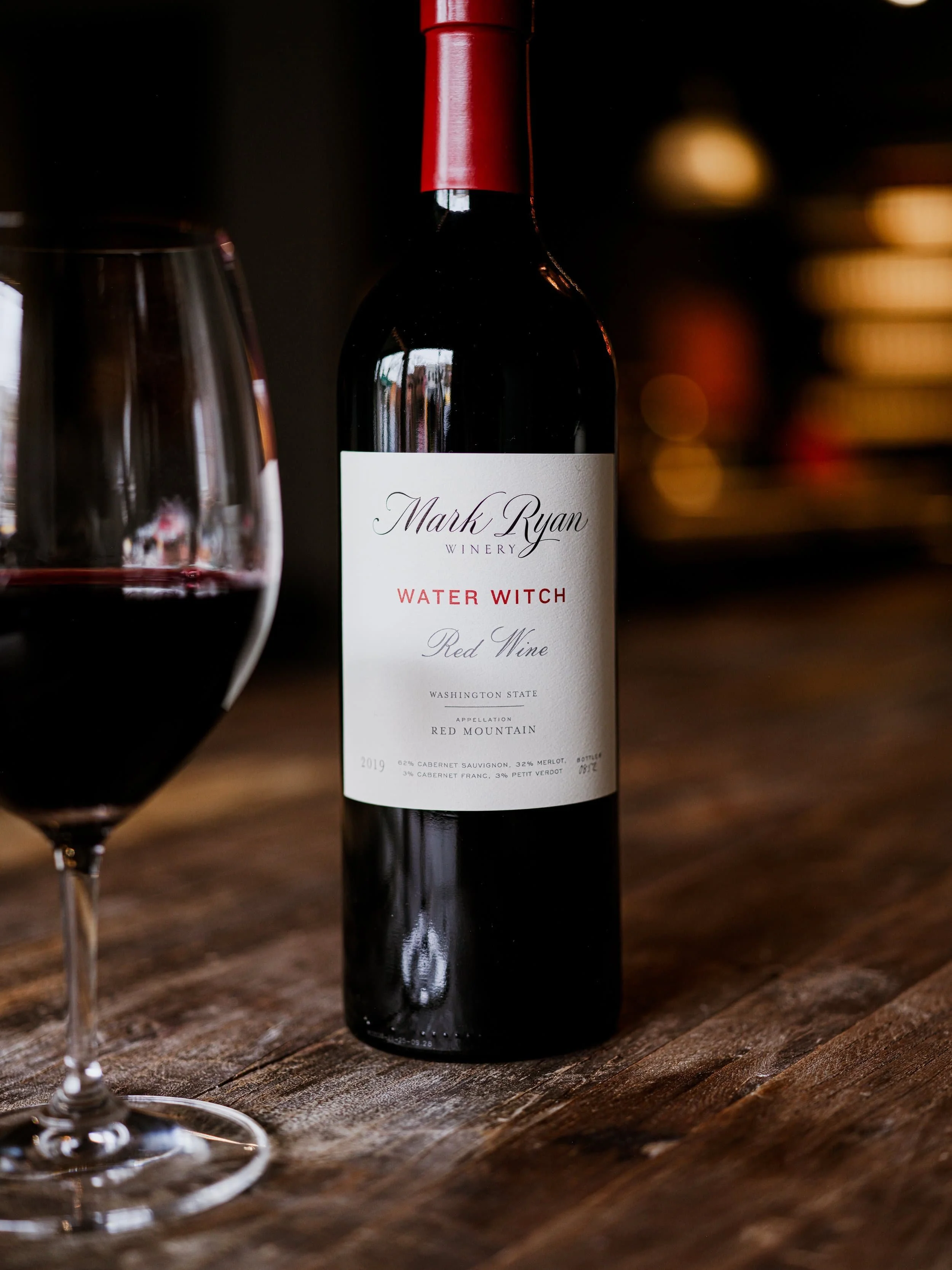 A bottle of Mark Ryan Water Witch on a wooden table next to a glass of red wine.