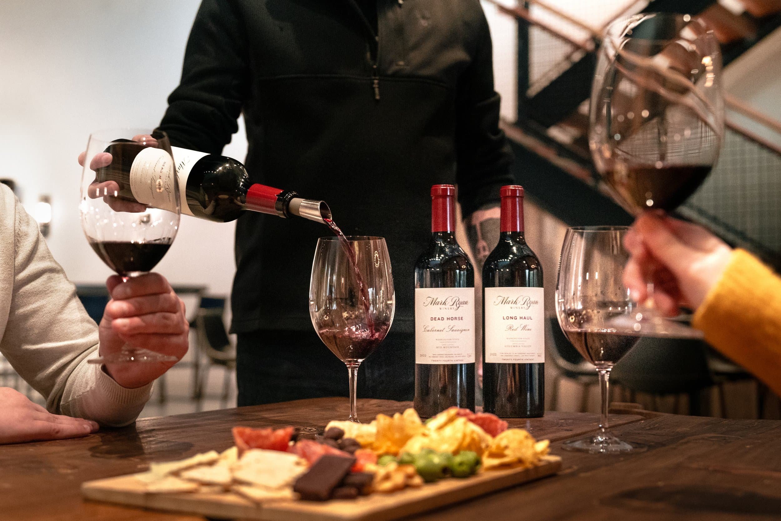 People enjoying red wine at the Mark Ryan Winery Woodinville tasting room with wine bottles, glasses, and a cheese platter on the table.