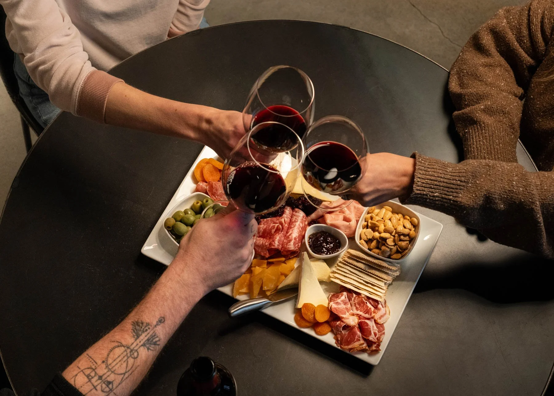 Three wine glasses clinking together in a toast above a beautifully arranged charcuterie board featuring cured meats, artisan cheeses.