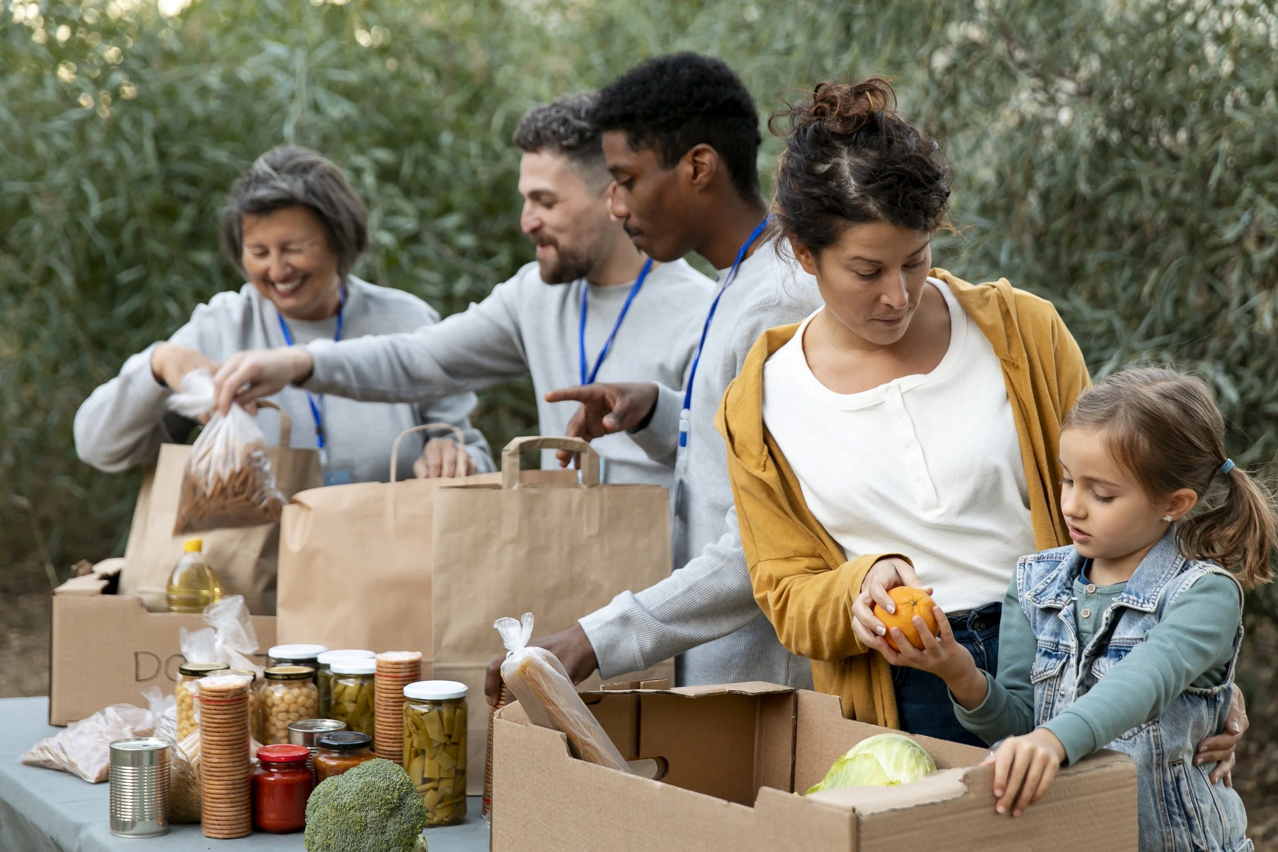 Group of people at a food collection or donation event outdoors, with shelves of canned goods, jars, and produce like broccoli, interacting happily.