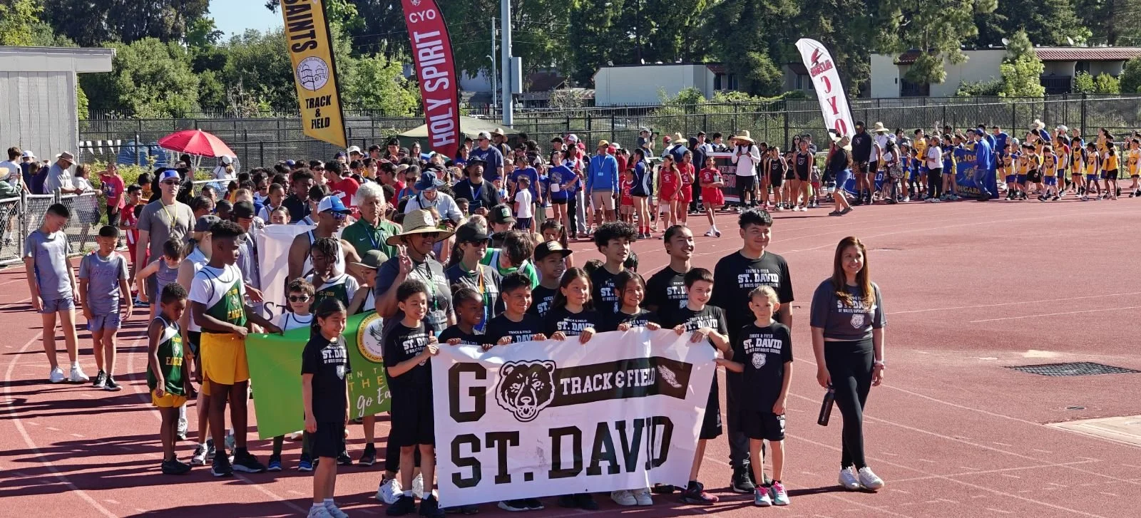 A large group of children and adults gathered in a track and field area, with some holding banners, wearing athletic clothing, and participating in a community or school track event on a sunny day.