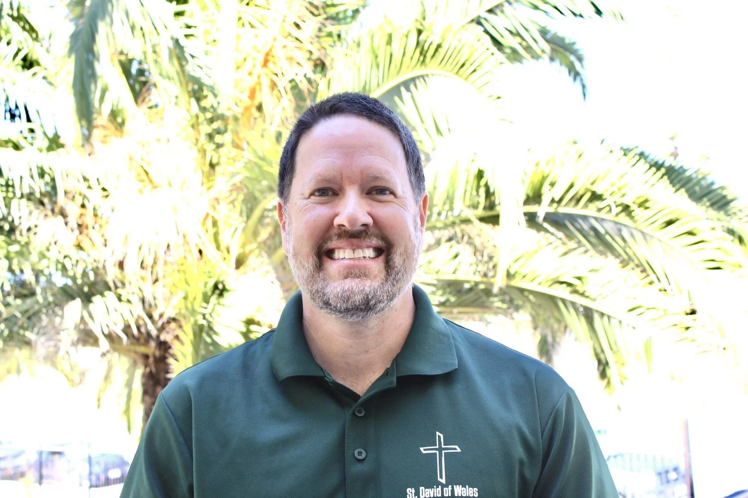 A smiling man with a beard and short dark hair, wearing a dark green polo shirt with a cross and the words "St. David of Wales" on it, standing outdoors with palm trees in the background.