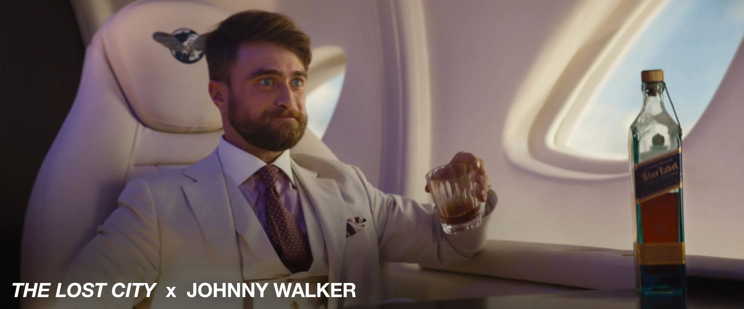 A man with a beard and mustache, wearing a white suit, purple shirt, and patterned tie, sitting in an airplane seat with a serious expression and holding a glass of whiskey. There is a bottle of whiskey on the tray table beside him, and an airplane w
