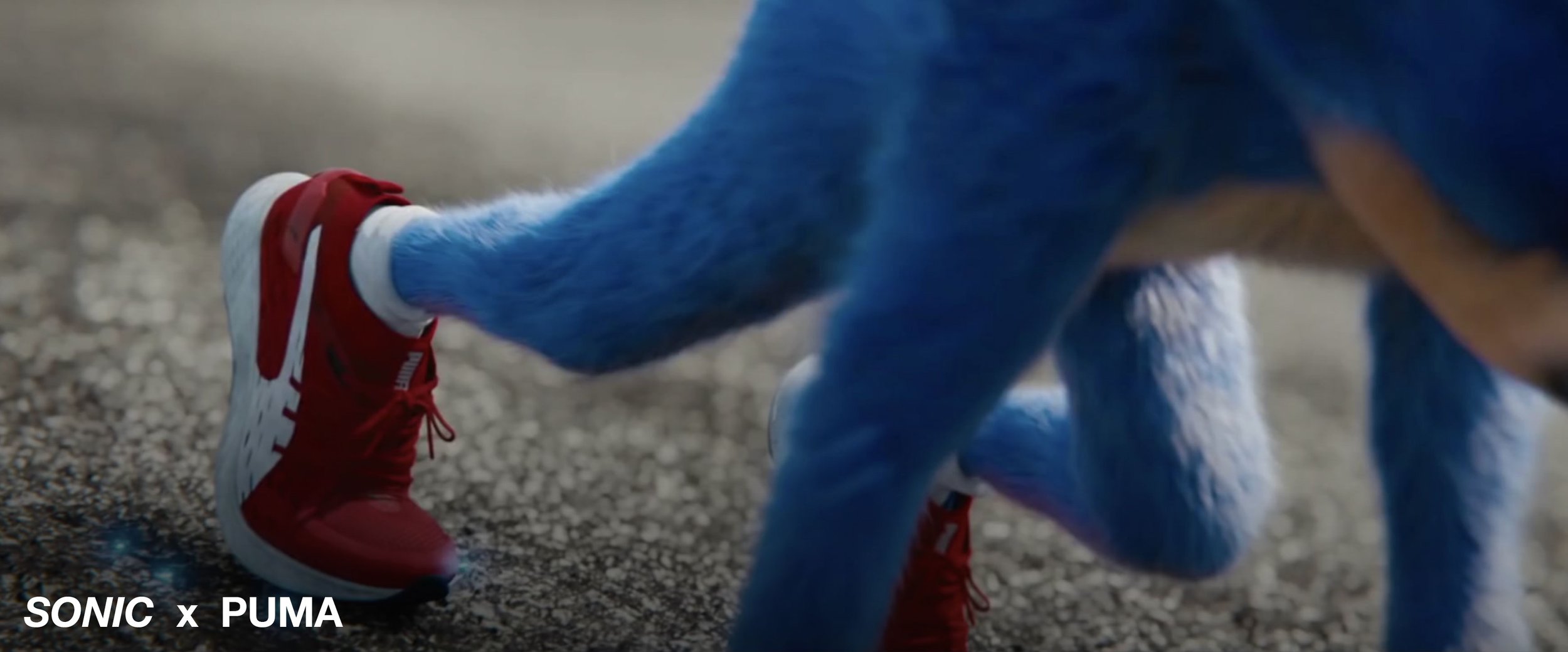 Close-up of a person's foot wearing a red Puma sneaker on a textured ground, with a blue furry creature's arm reaching towards the shoe, part of a Sonic the Hedgehog costume, in the background.