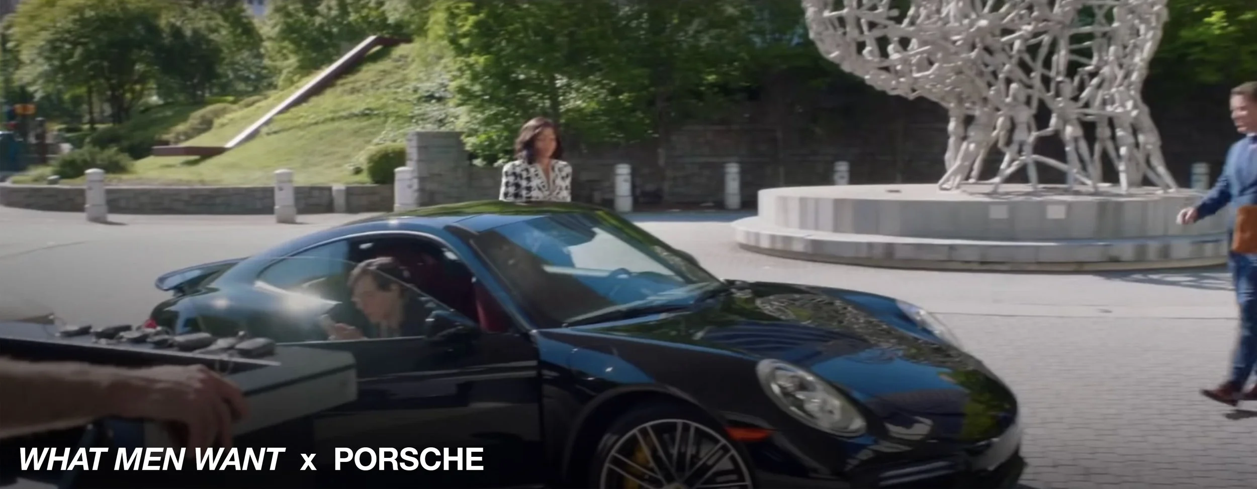 A black Porsche sports car is parked on a city street near a decorative sculpture; two women and a man are nearby, and a DJ is playing music from a turntable in the foreground.