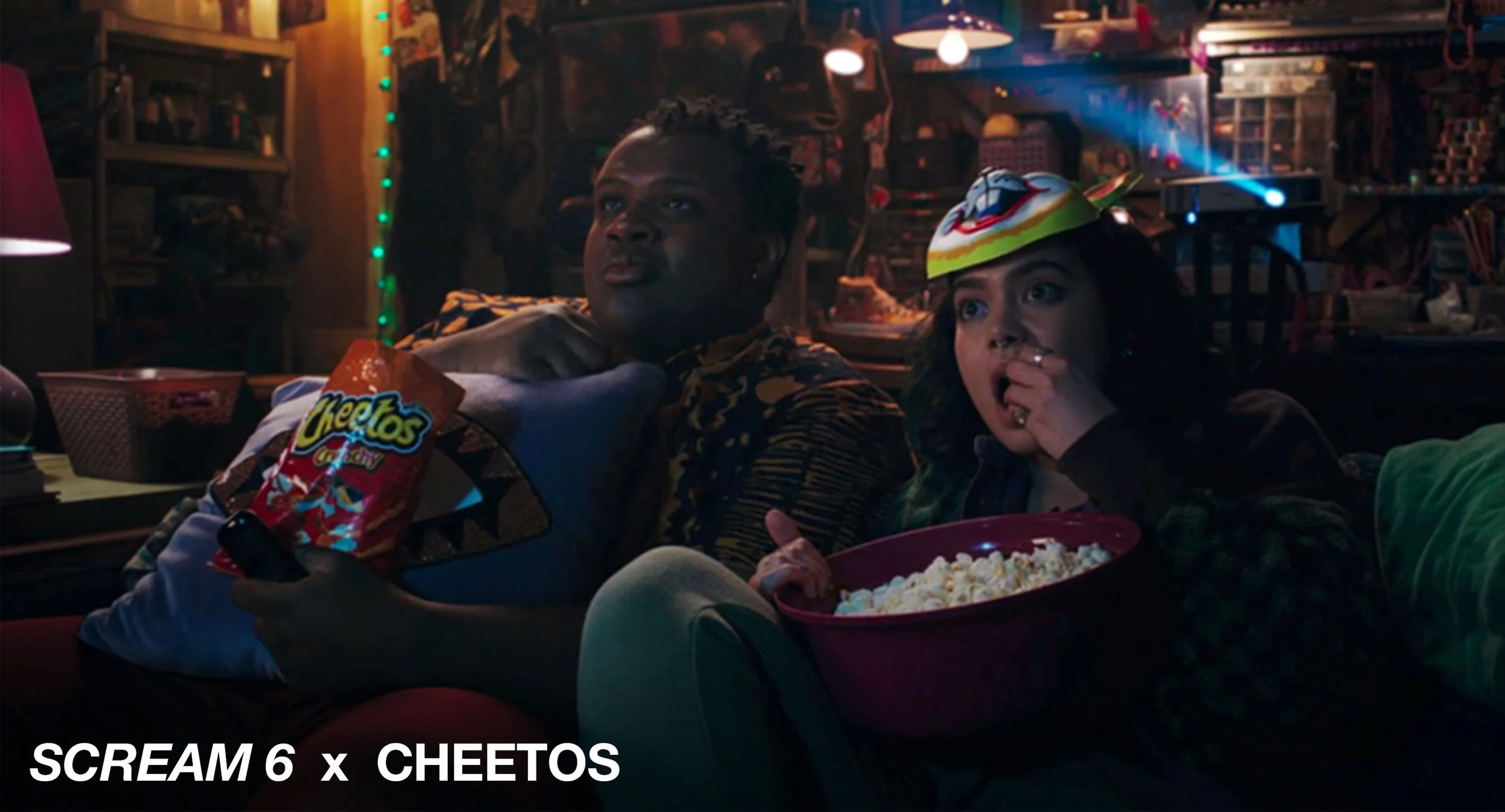 Two people sitting on a couch watching TV in a dimly lit room decorated with string lights. One is holding a bag of Cheetos and a remote, and the other has a large bowl of popcorn. The person on the right is wearing a colorful mask or hat.