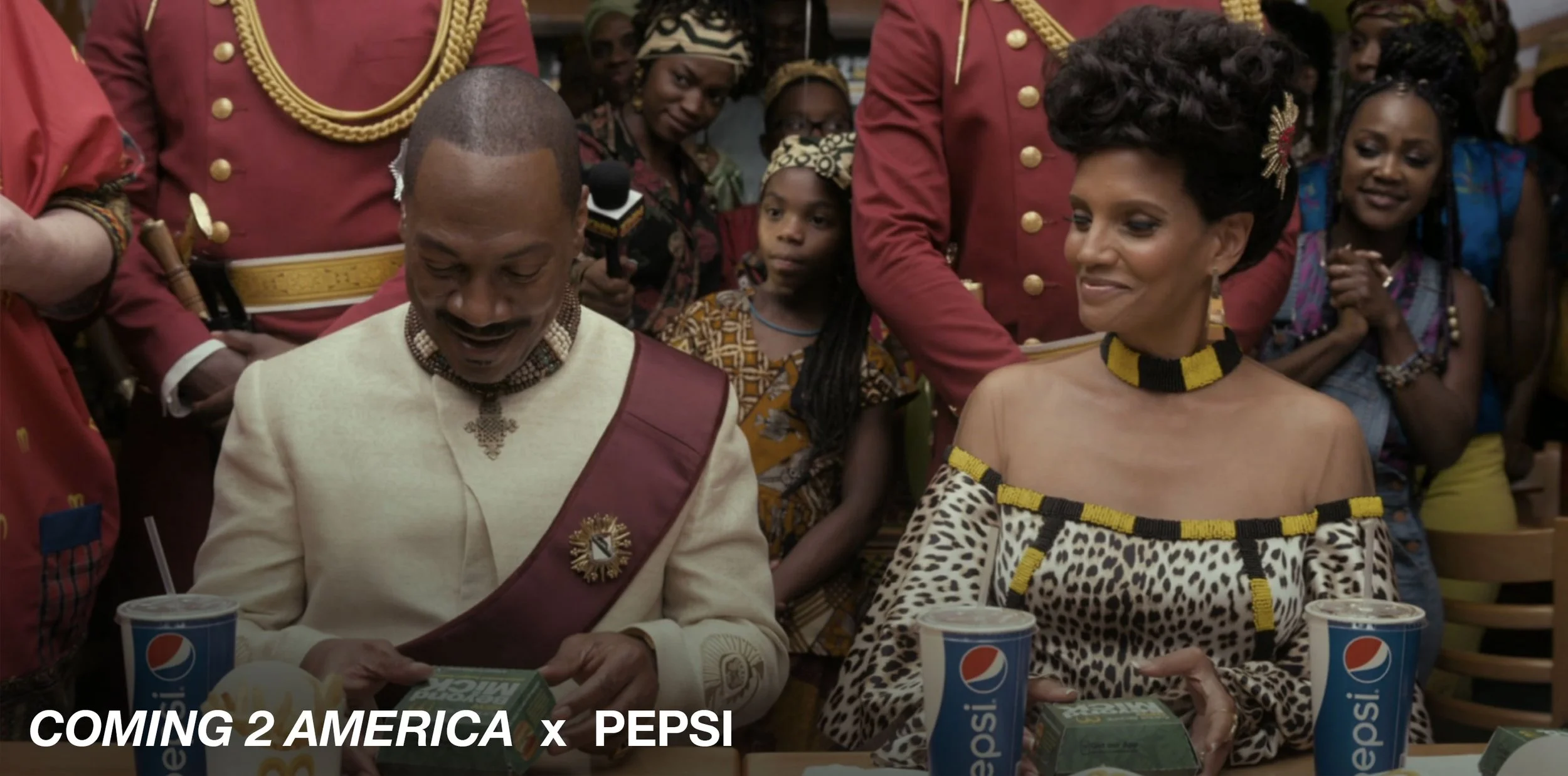 A man and woman sitting at a table with Pepsi cups and food, surrounded by people dressed in traditional African clothing, at an event celebrating African culture.