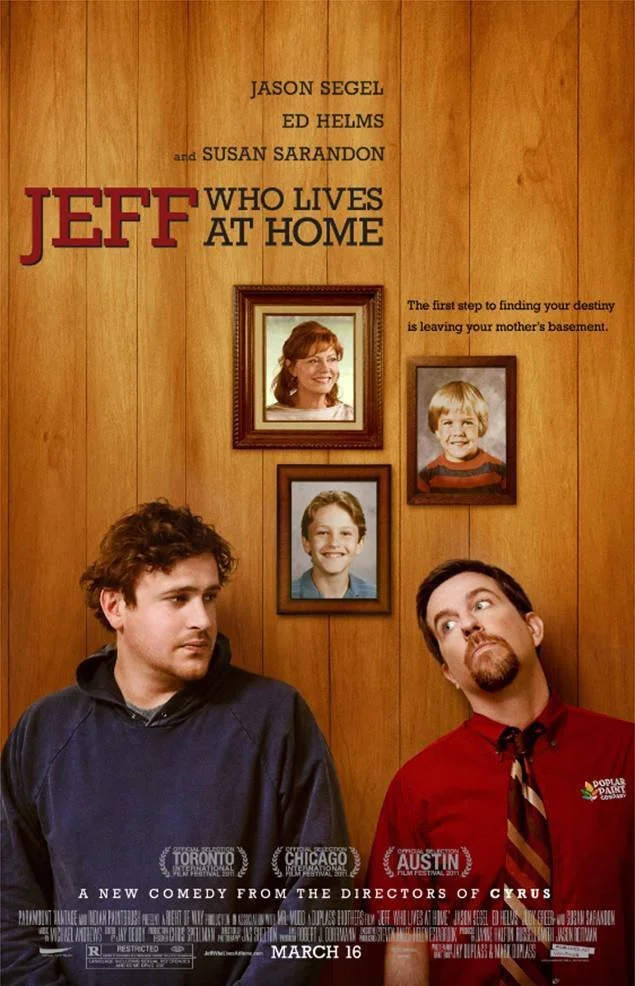 A movie poster for 'Jeff Who Lives at Home' featuring two men; one with curly hair wearing a dark hoodie, and another with straight hair and a goatee wearing a red shirt with a tie. The background is a wooden wall with three framed childhood photos o