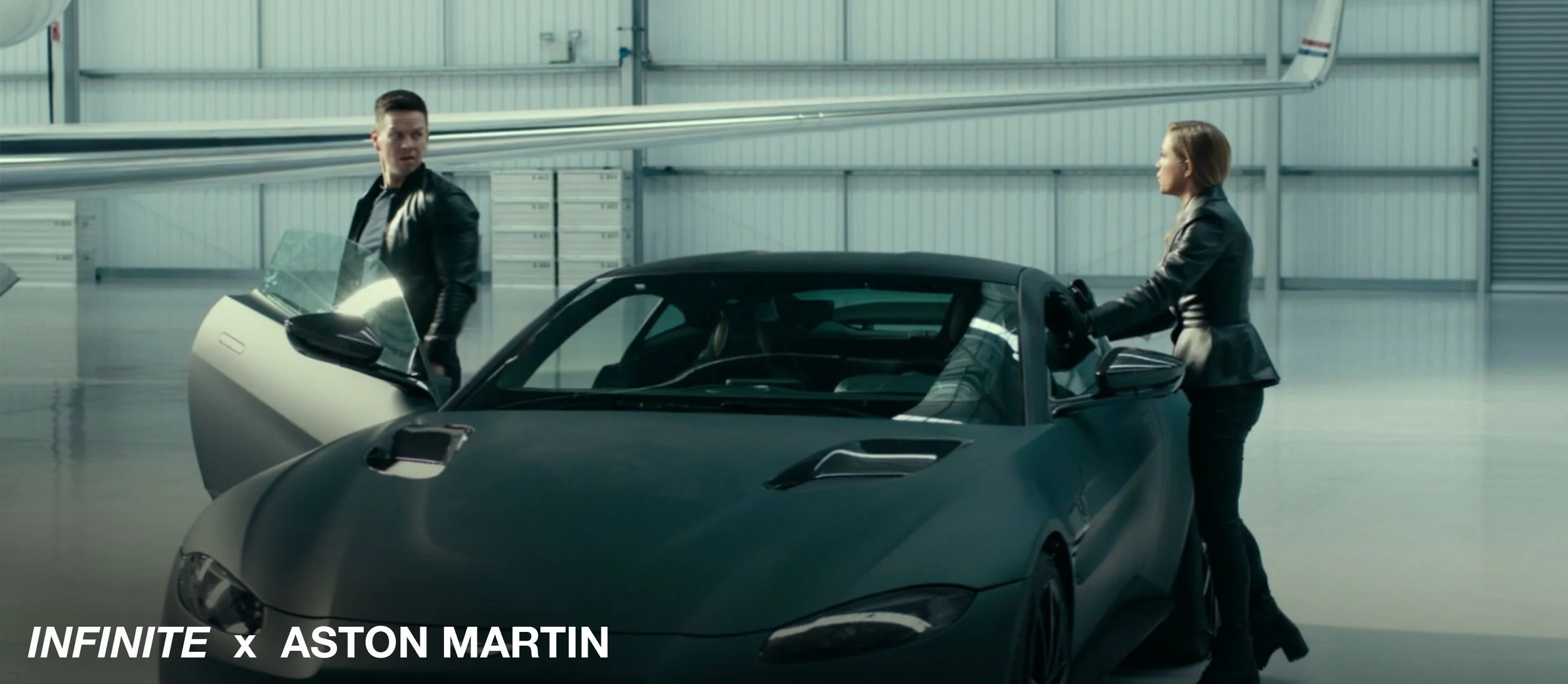 A man and woman are standing next to a matte black luxury sports car in an indoor garage or hangar, with the woman reaching into the car and the man looking at her. The setting is spacious and industrial with metal walls and ceiling.