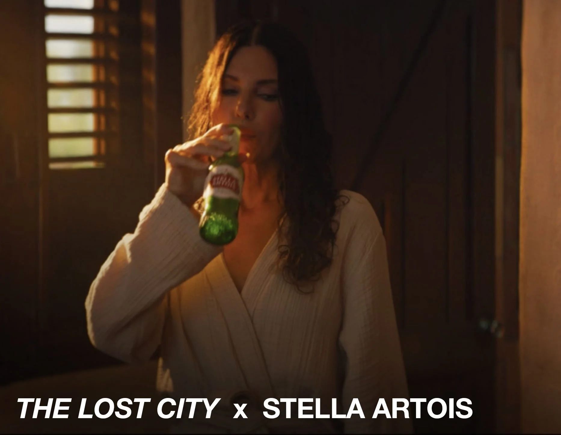 A woman with dark hair and a cream-colored robe drinking a Stella Artois beer in a dimly lit room.