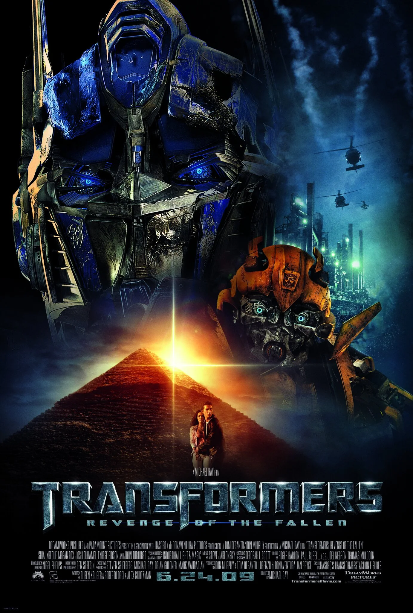 Movie poster for Transformers: Revenge of the Fallen featuring large robotic characters with glowing eyes, an industrial cityscape with helicopters, pyramids with a bright light, and two human characters standing in front.