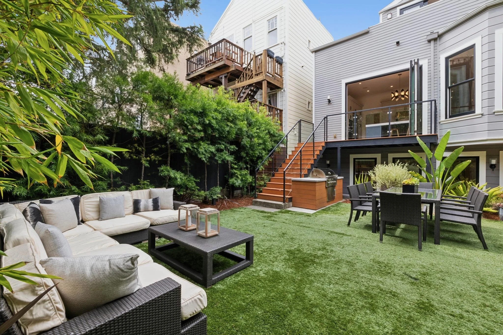 A backyard patio with outdoor furniture, including a white cushioned sofa with throw pillows, a black wicker coffee table with lanterns, and a dining table with black chairs. The area has artificial grass and is surrounded by lush green plants and tr