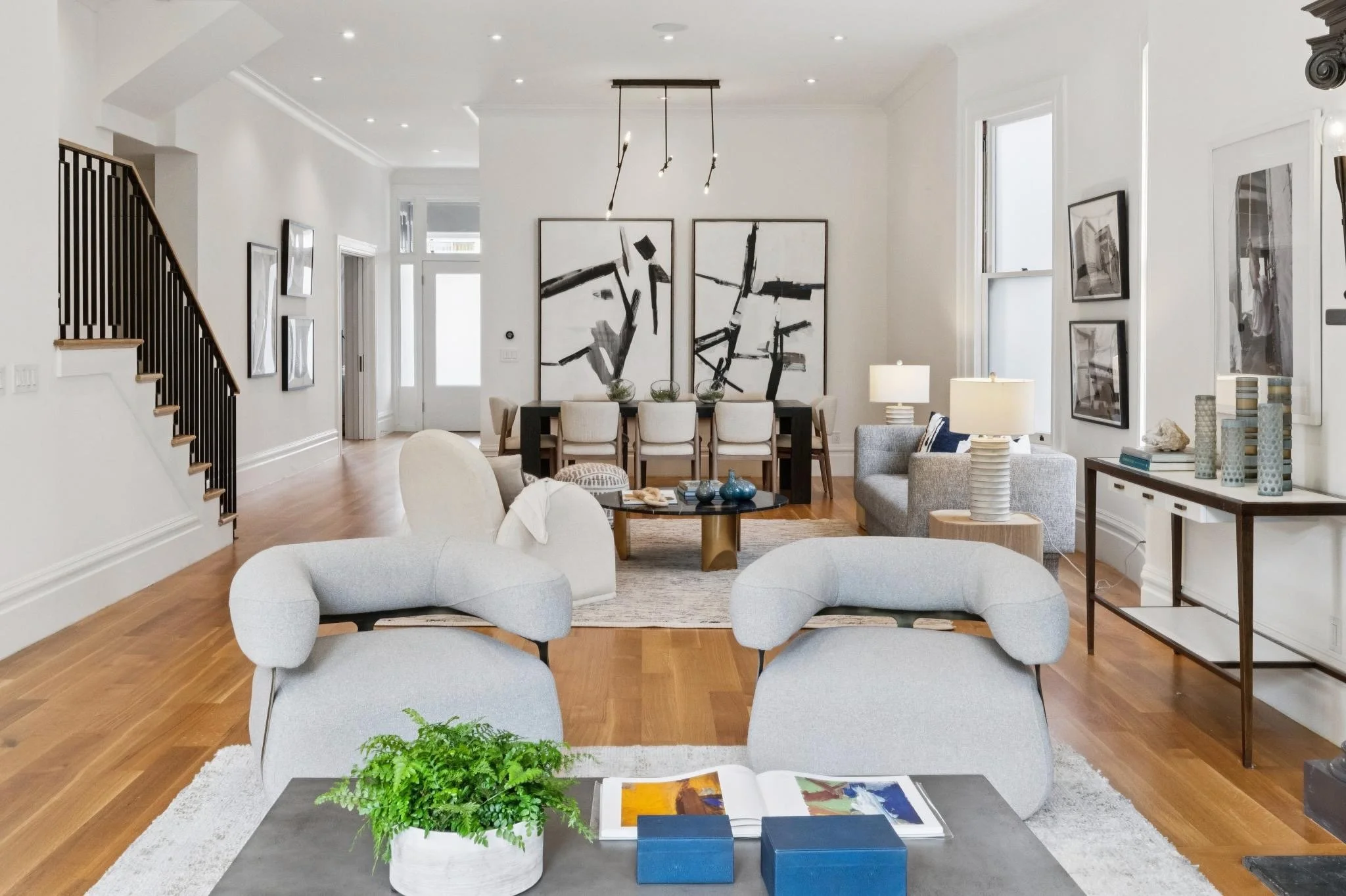 Living room with white walls, hardwood floors, modern furniture, black and white abstract art, and natural light from tall windows.