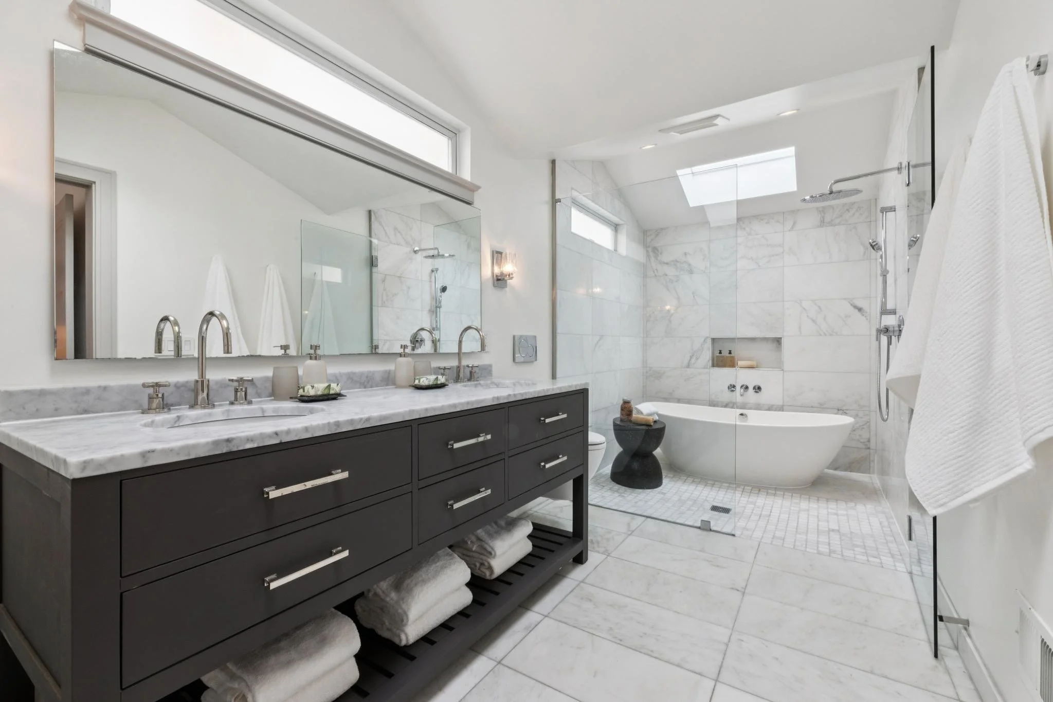 Modern bathroom with vanity, marble countertop, large mirror, shower area, bathtub, and white towels.