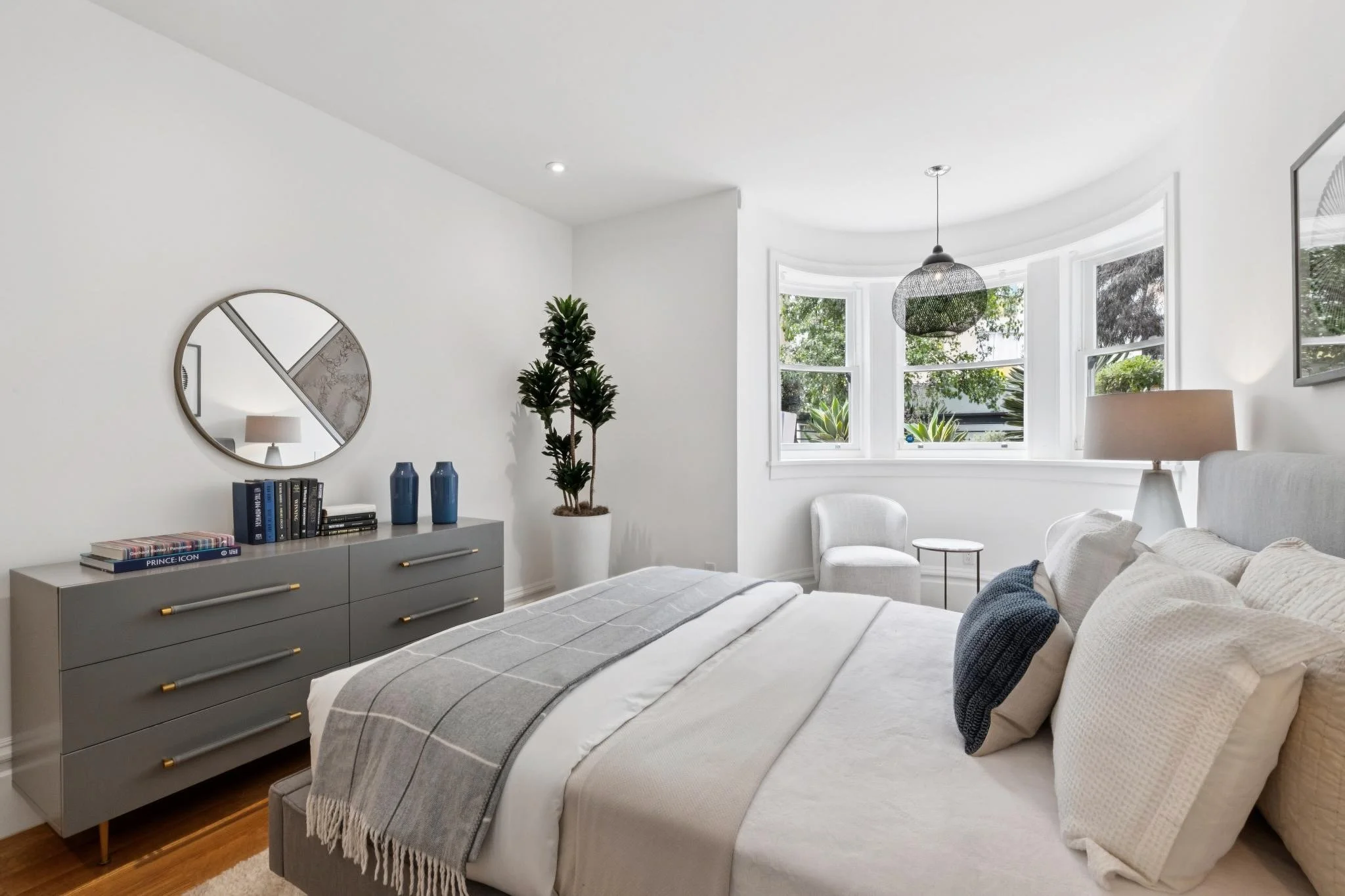 A modern bedroom with a bed covered in a gray and white striped blanket, a bedside lamp, a gray dresser with books and decorative blue vases, a tall potted plant, a white armchair, and a window with a view of green trees outside.