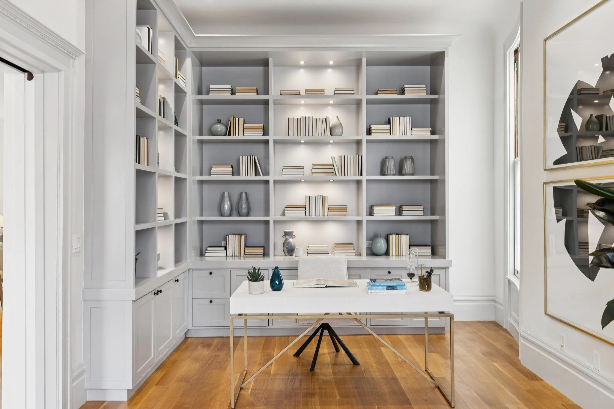 A modern white office with built-in bookshelves filled with books and decorative items, a white desk with a chair, glass vases, and a small potted plant, with wood flooring and large windows.