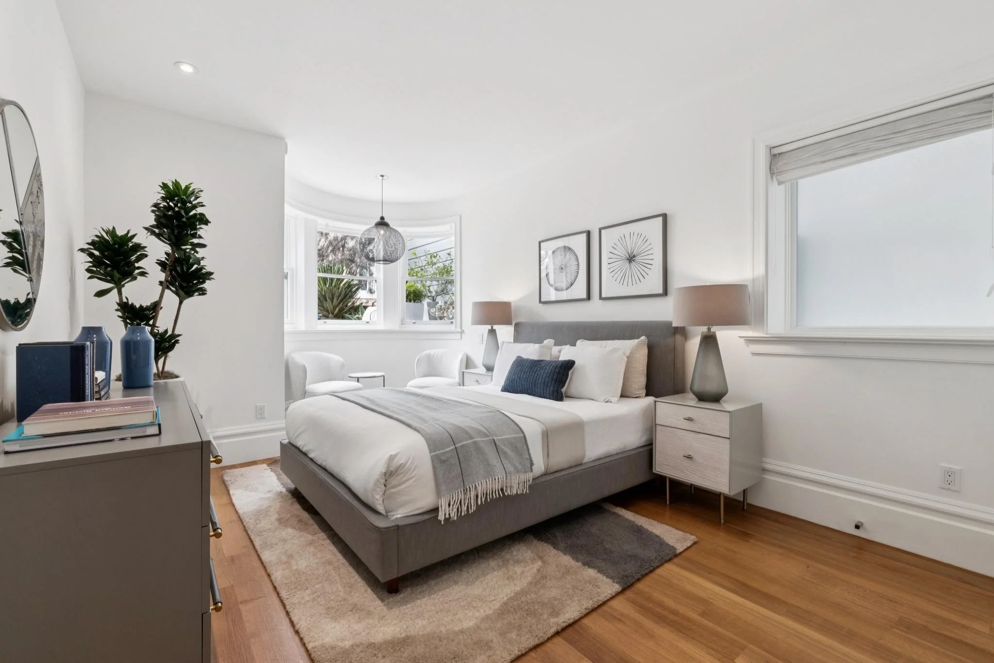 A bright, modern bedroom with a large bed, white linens, a gray headboard, and two beige nightstands with lamps on each side. There are two framed abstract art pieces above the bed, a window with a view of plants outside, and a gray dresser with book