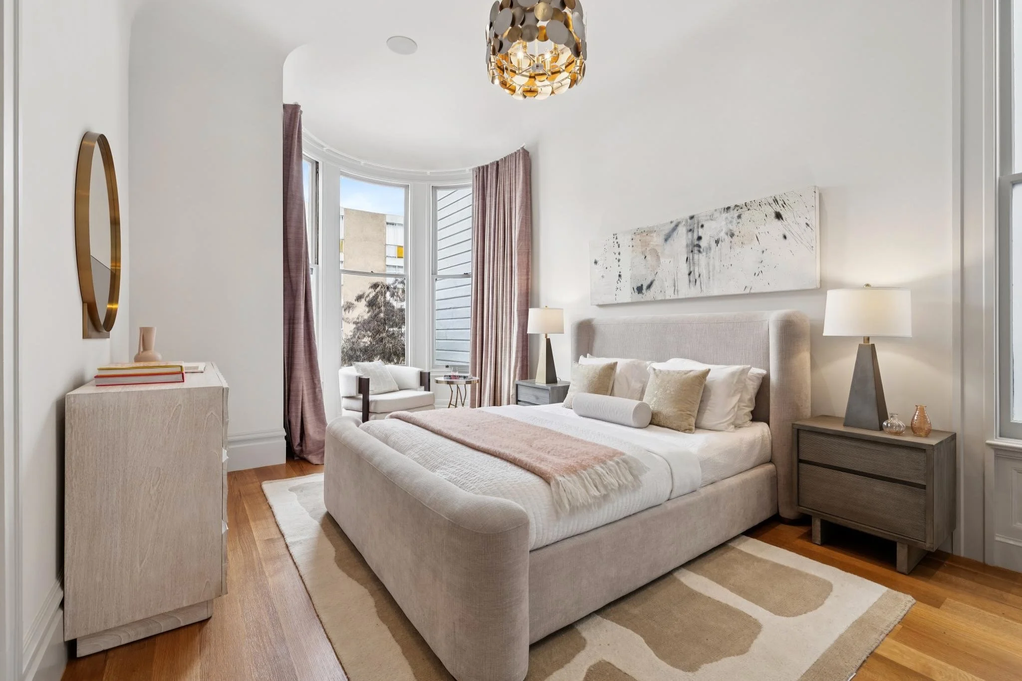 A modern bedroom with a large bed, beige upholstered headboard, and neutral linens. There are two bedside tables with lamps, an abstract painting above the bed, and a seating area near a bay window with pink curtains. The room has hardwood floors and