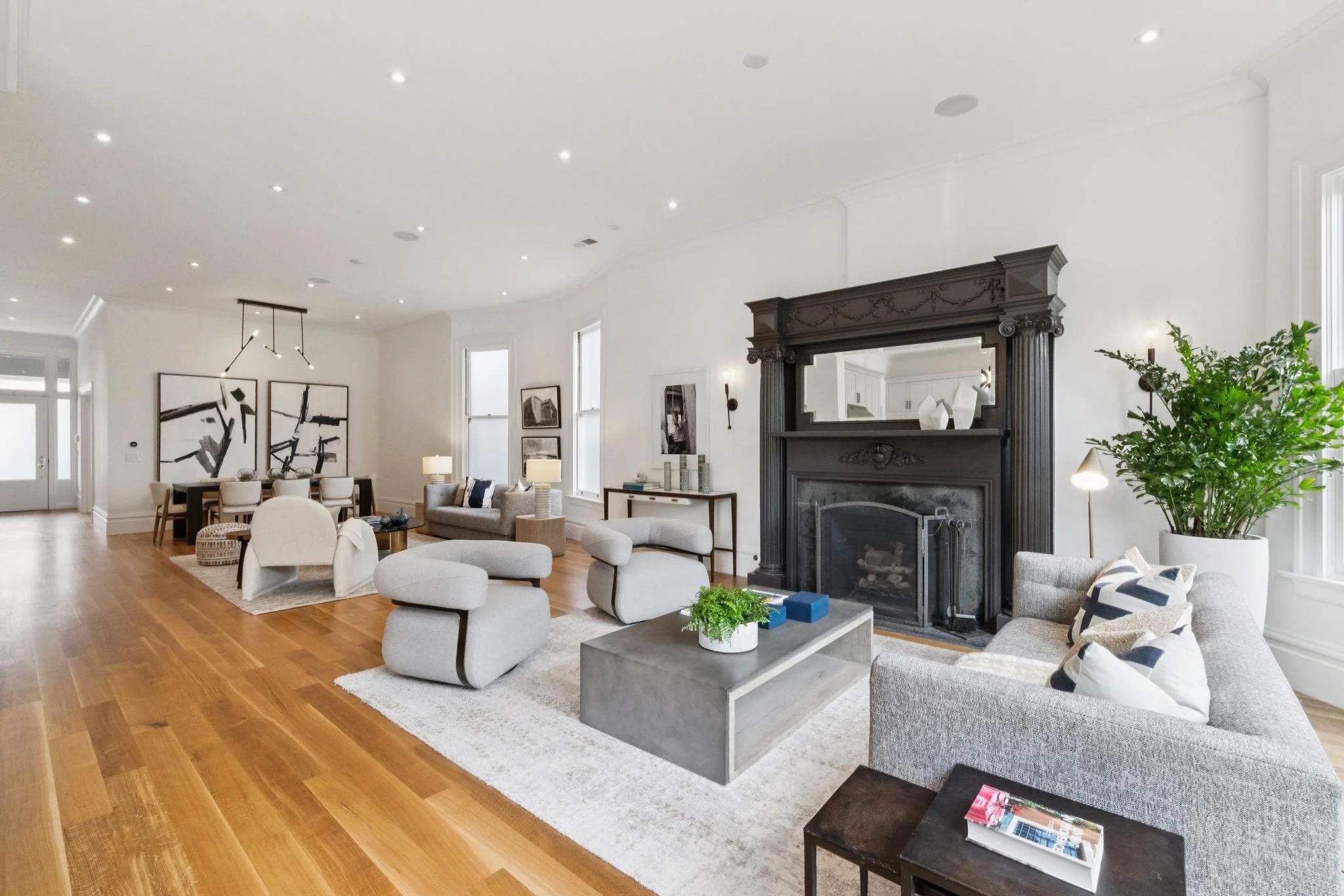 A spacious living room with white walls, wooden flooring, and modern furniture. There are several armchairs, a gray sofa, and a coffee table. A large ornate black fireplace with a mirror above it is on one wall, and the room is decorated with black-a
