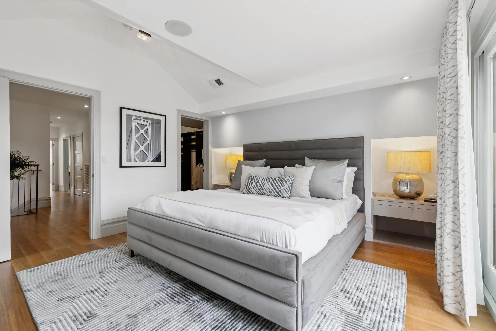 A modern bedroom with a plush gray bed, white bedding, decorative pillows, wooden nightstand with lamp, and a large window with white patterned curtains.