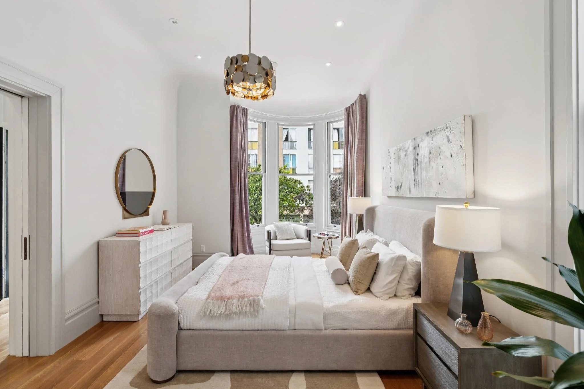 Bright, modern bedroom with a white upholstered bed, neutral decor, a large window with purple curtains, and a gold and white pendant light fixture.