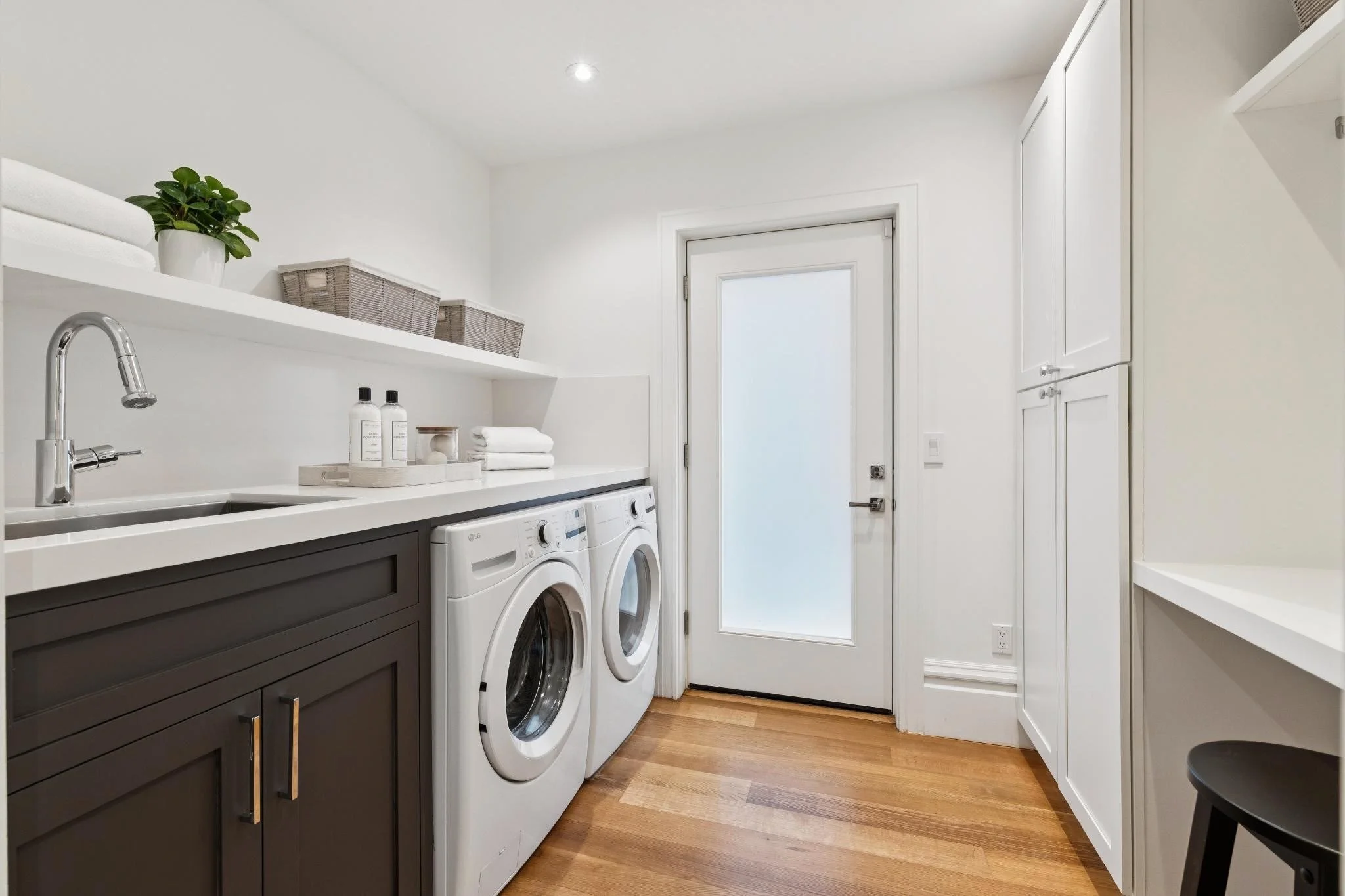 A laundry room with white walls, a door with a frosted window, a white shelf with a potted plant and baskets, a dark-colored cabinet with gold handles, two front-loading washers, and a white cabinet on the right side.